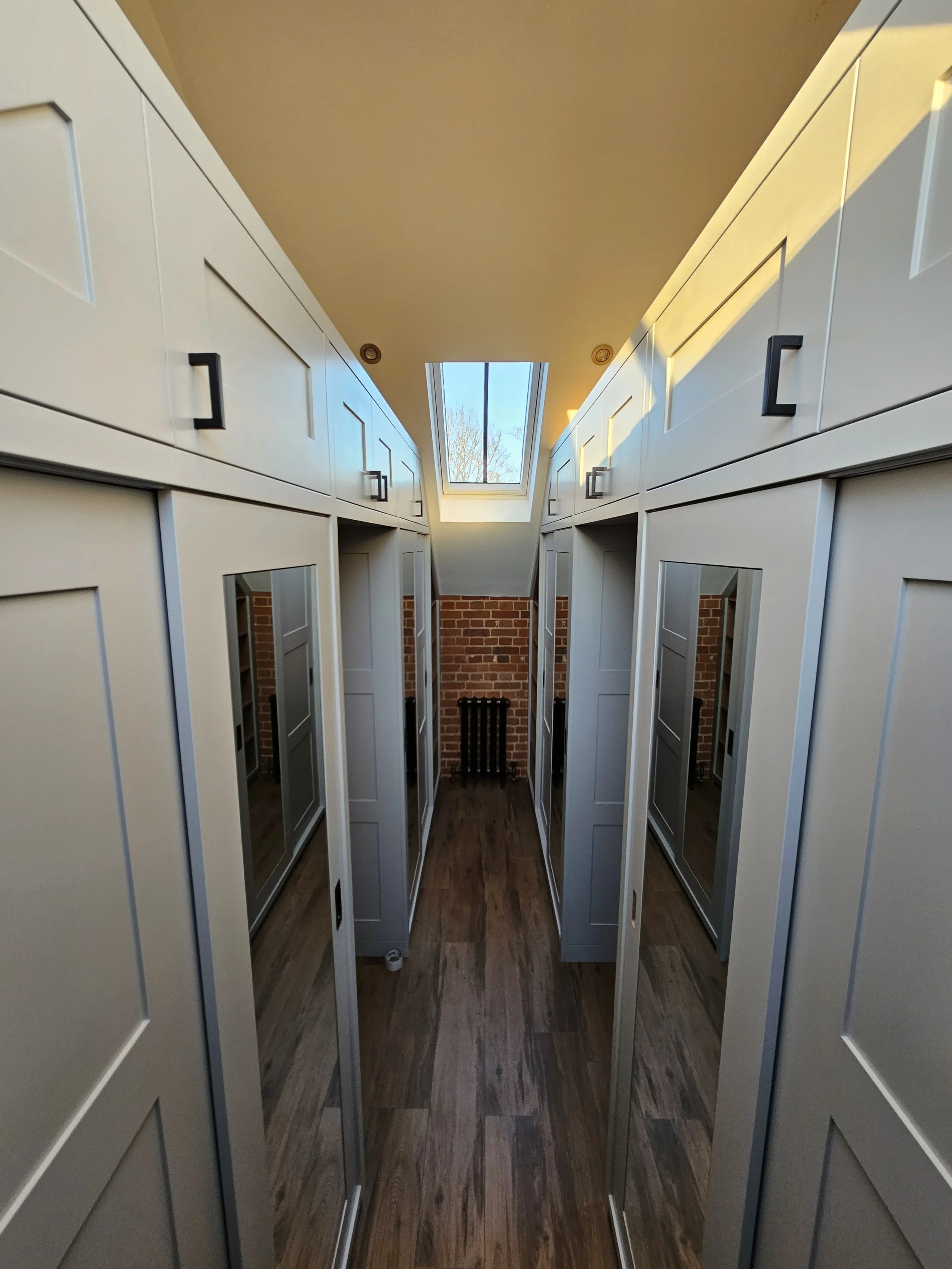 A walk-in closet with white cabinetry, mirrored closet doors, wood flooring, a skylight, and brick wall at the far end.