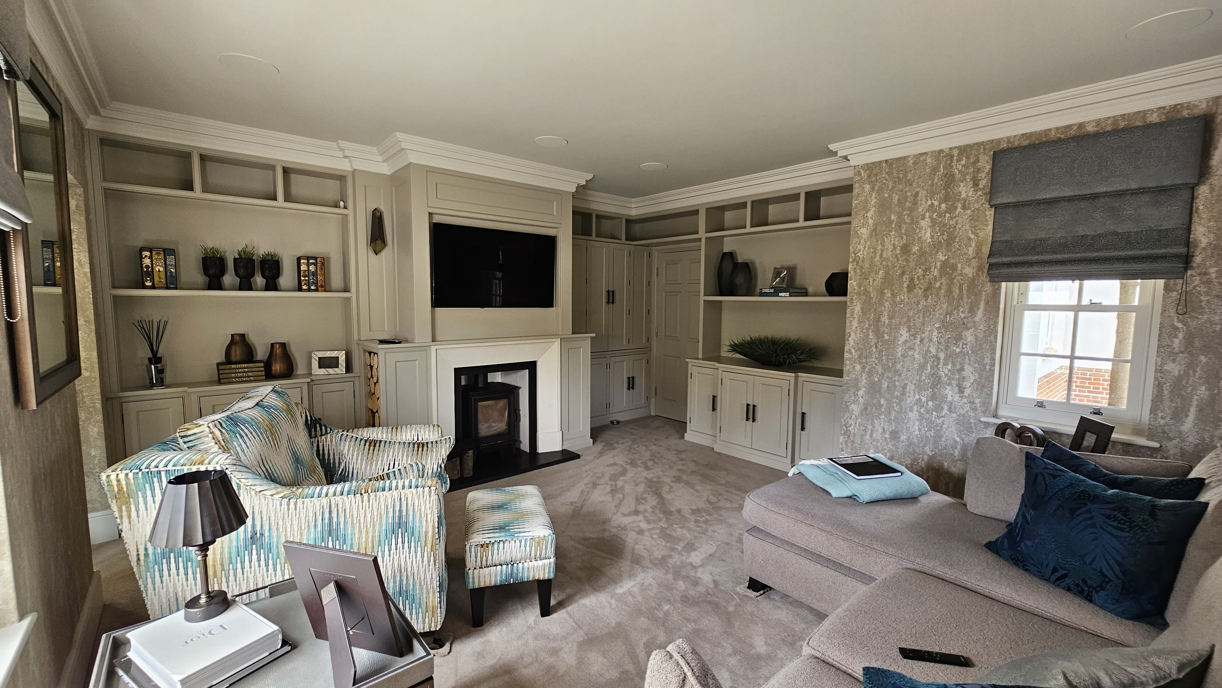 Living room with built-in shelves, fireplace, gray sectional sofa, armchair with colorful pattern, window with gray window treatment, and various decorations such as vases, books, and pillows.