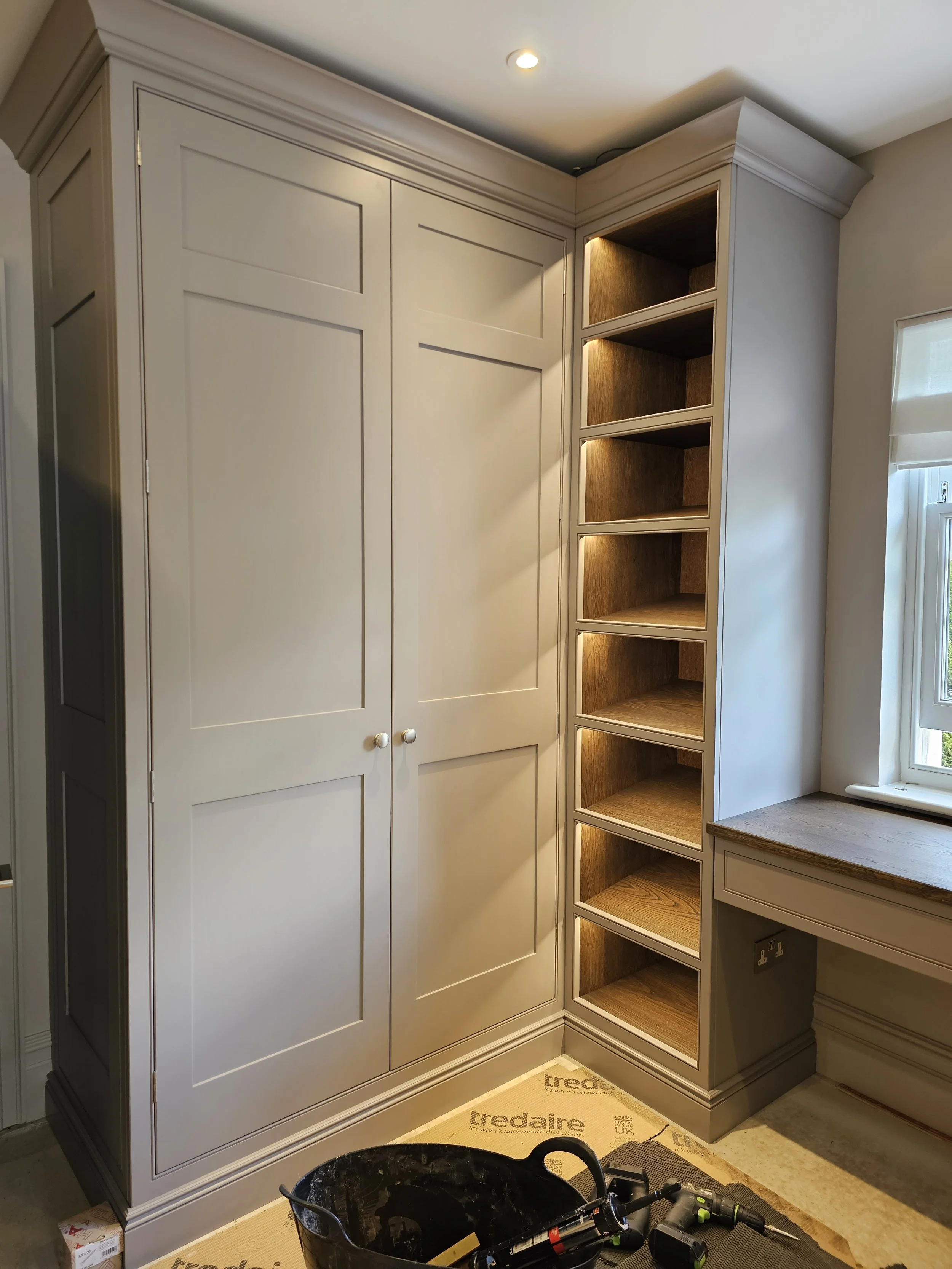 Built-in white wardrobe with two doors and a corner open shelving unit with five wooden shelves and integrated lighting, next to a window with a window seat.