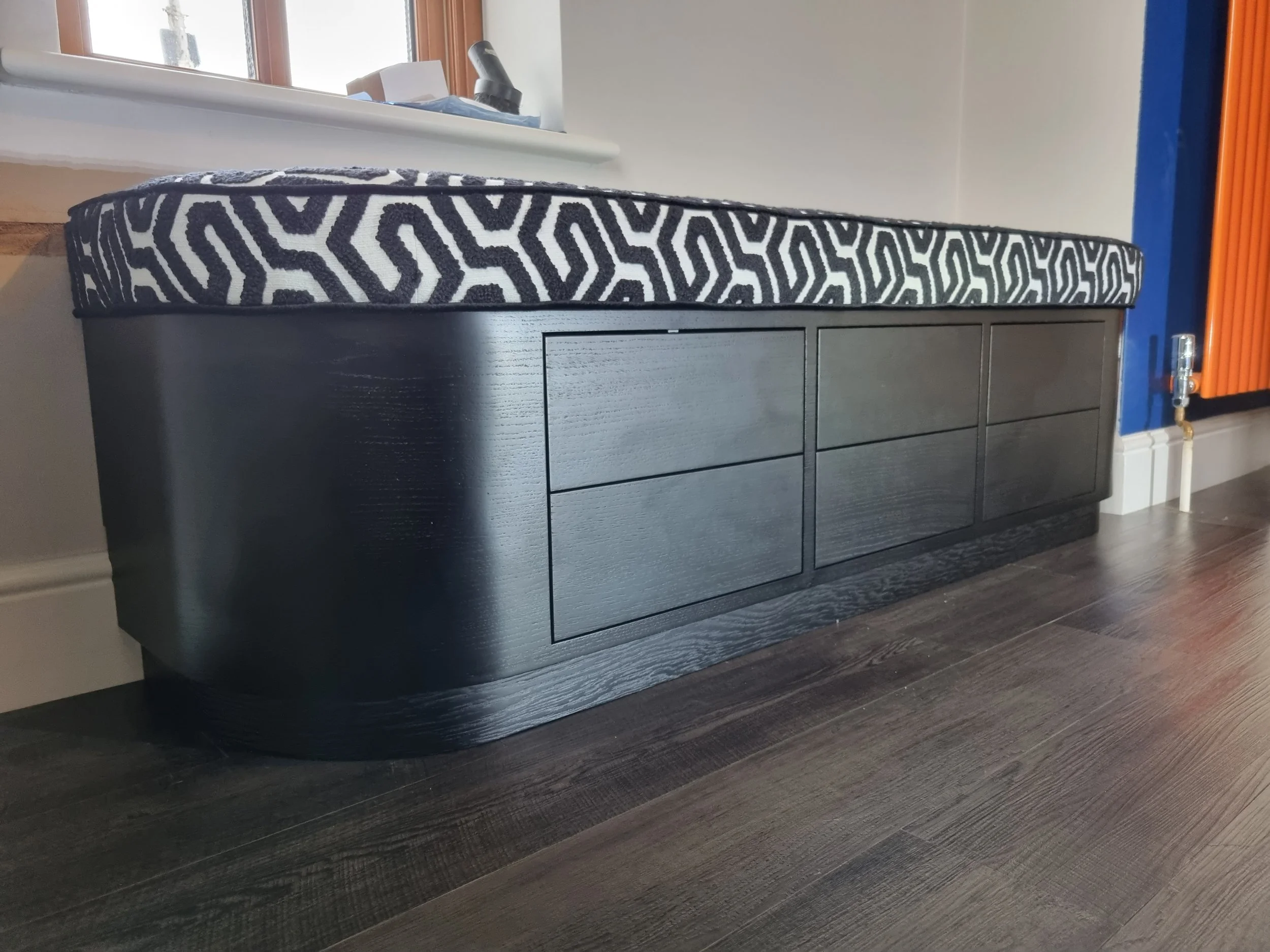 Black wooden storage bench with six drawers under a window with a patterned cushion on top.