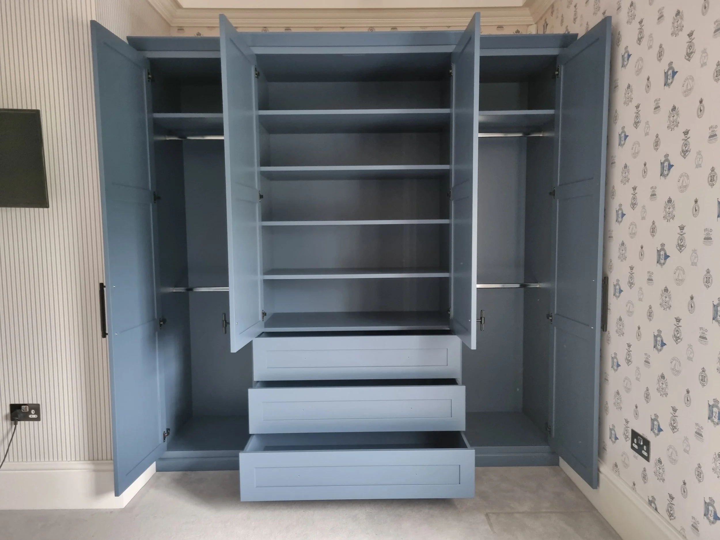 A large light blue wardrobe with open twelve compartments, some with hanging rods and others with shelves, and four drawers at the bottom. It is situated in a room with wallpaper featuring various blue and gray emblems on the right wall.