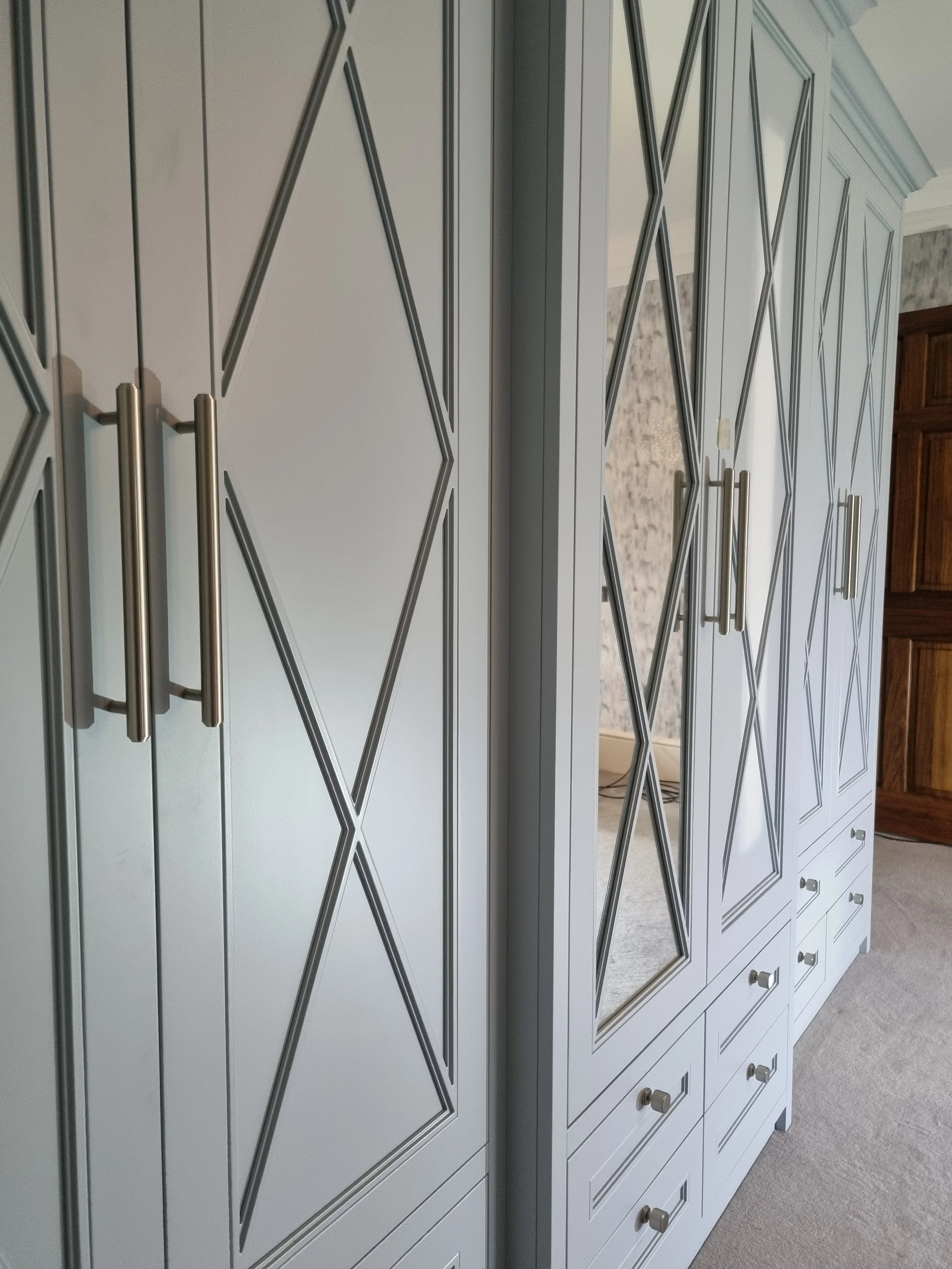 White built-in wardrobe with diamond-patterned details and silver handles, reflected in a mirror, in a room with beige carpet and wooden furniture.