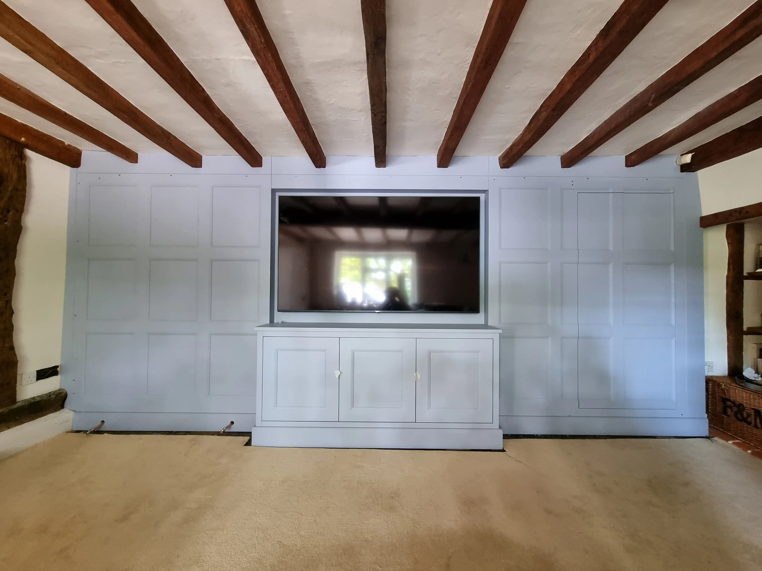 Living room with a large flat-screen TV mounted on a wall with decorative paneling, above a white cabinet, and exposed wooden ceiling beams.