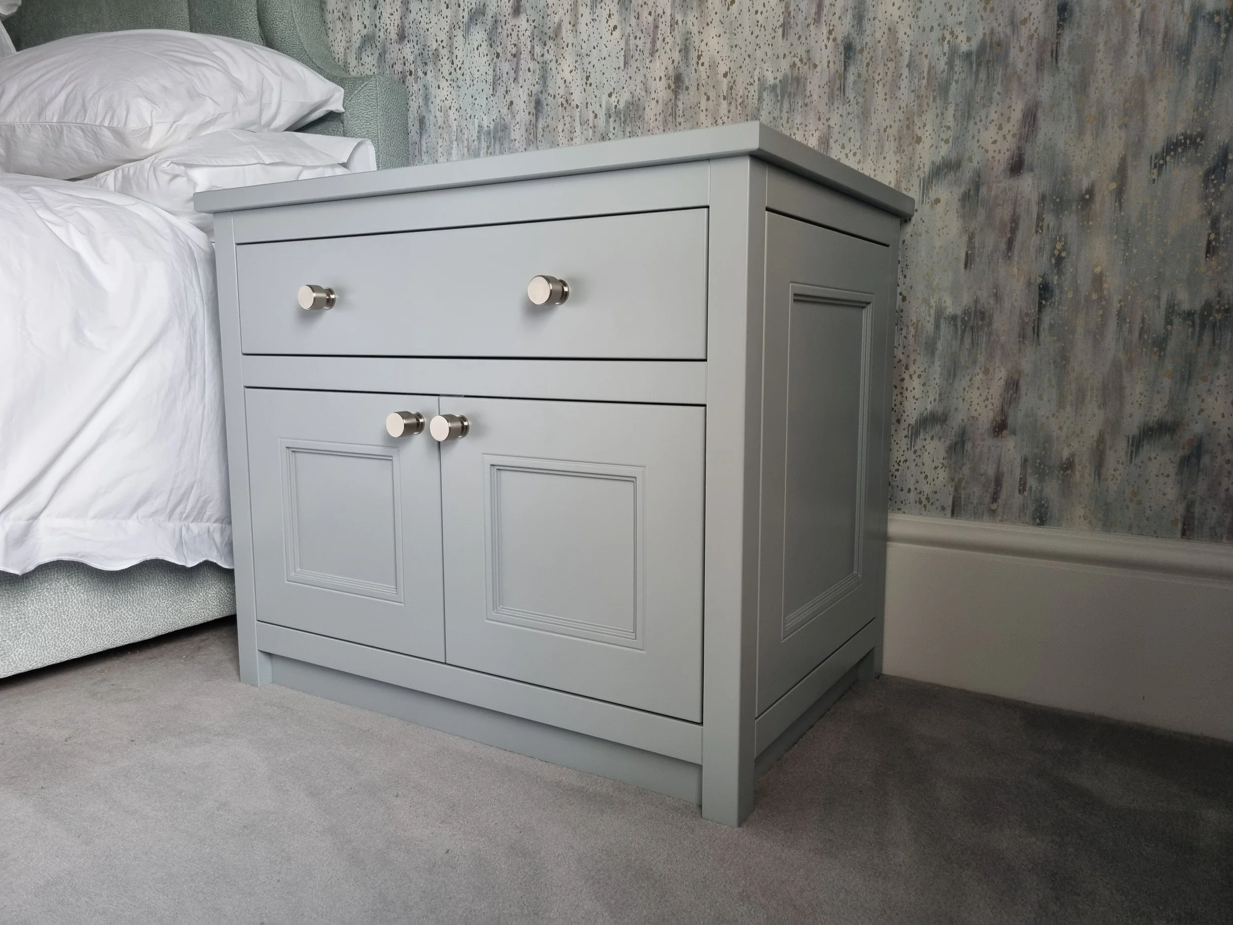 Light gray wooden nightstand with two drawers and two cabinet doors, placed beside a bed with white bedding, in a bedroom with patterned wallpaper and gray carpet.