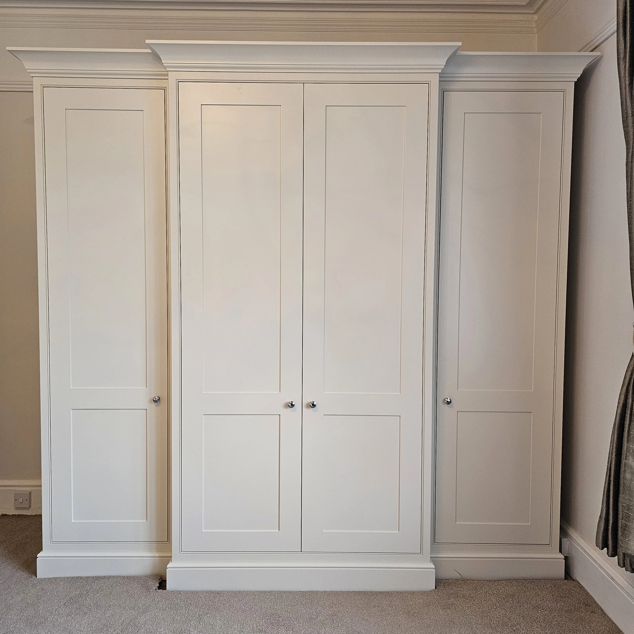 A large white wardrobe with four doors, two in the middle and one on each side, with decorative trim and small round knobs, situated in a room with beige carpet and curtains on the right.