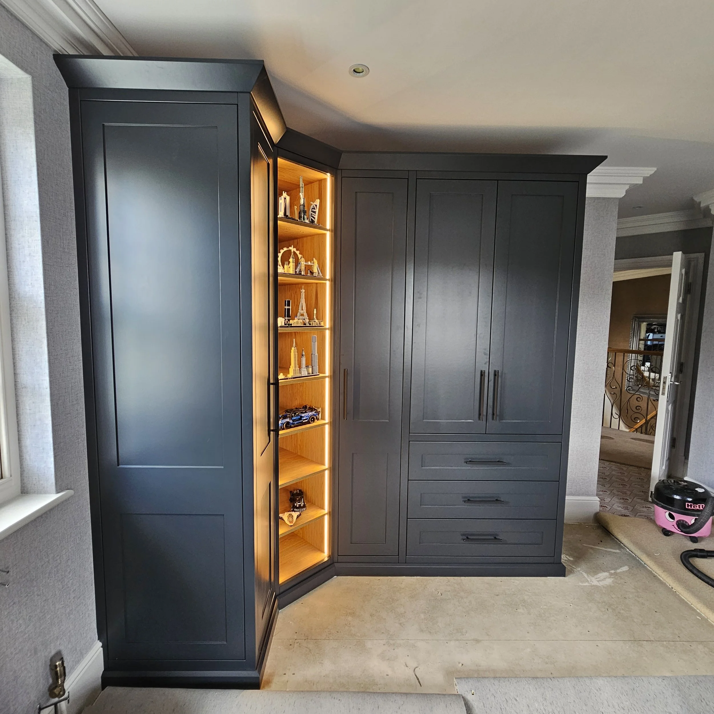 A large dark gray built-in cabinet with wood shelving on the left side, illuminated with warm lighting, displaying model souvenirs, with drawers and cabinet doors on the right side in a room with beige flooring and walls.