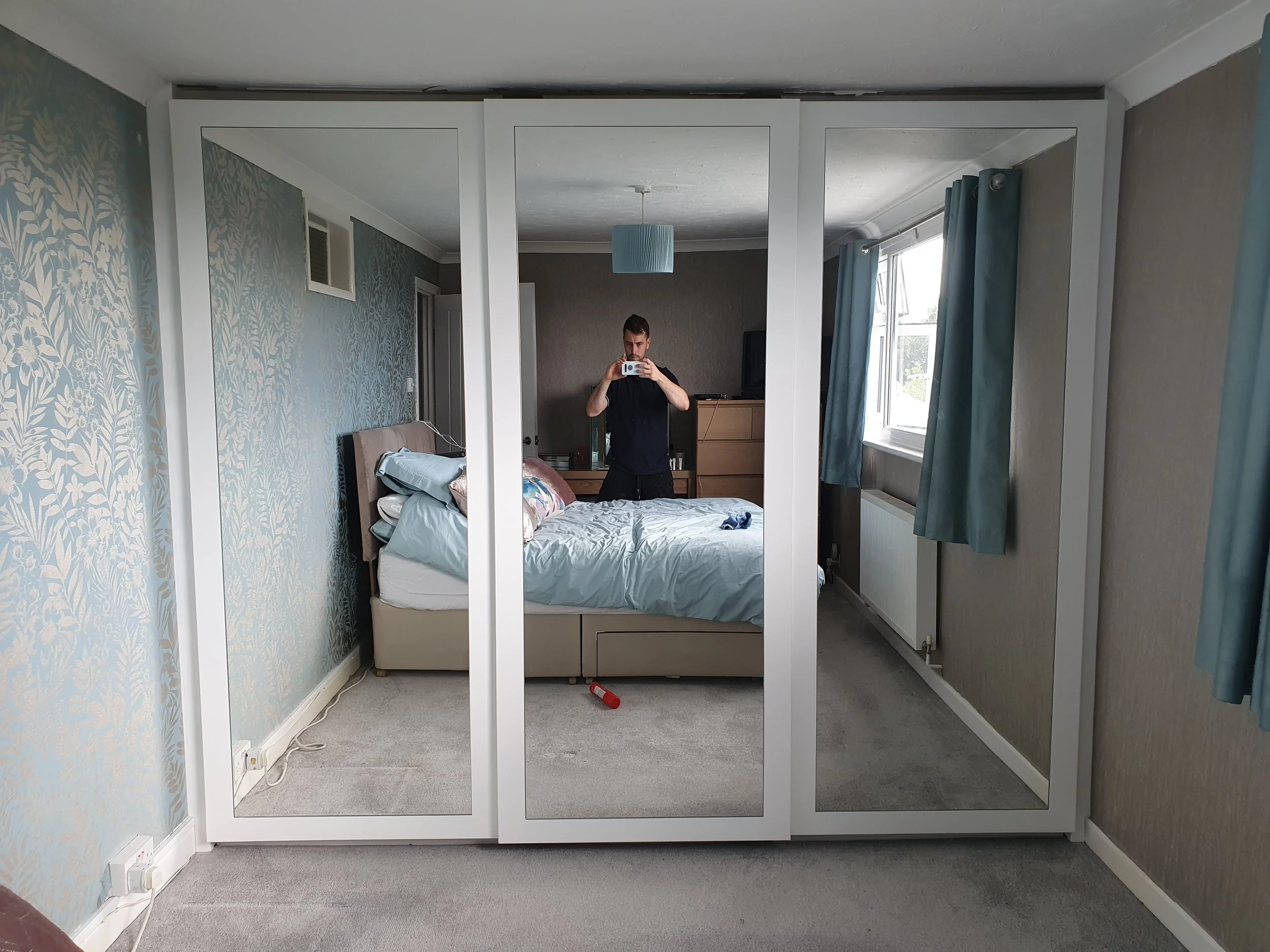 A man takes a selfie in a large bedroom with three mirrored closet doors. The room has light-colored walls, a window with blue curtains, a bed with disheveled bedding, and a grey carpeted floor.