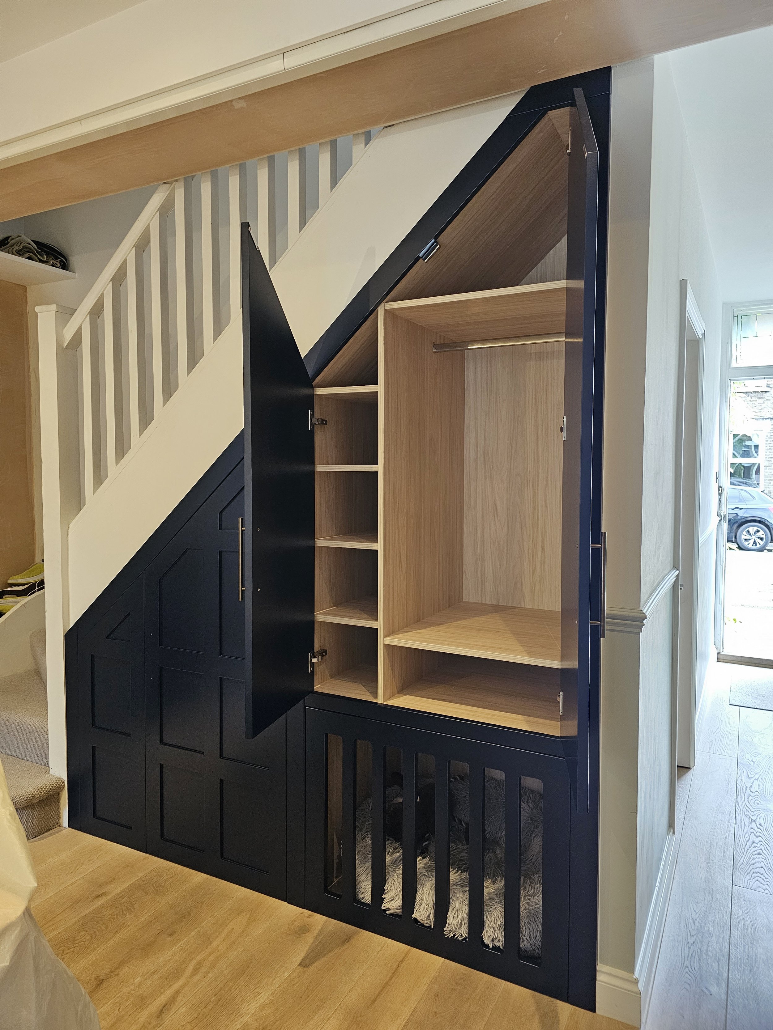 Built-in storage unit under staircase with black and wood finish, featuring open shelves and a dog bed underneath.