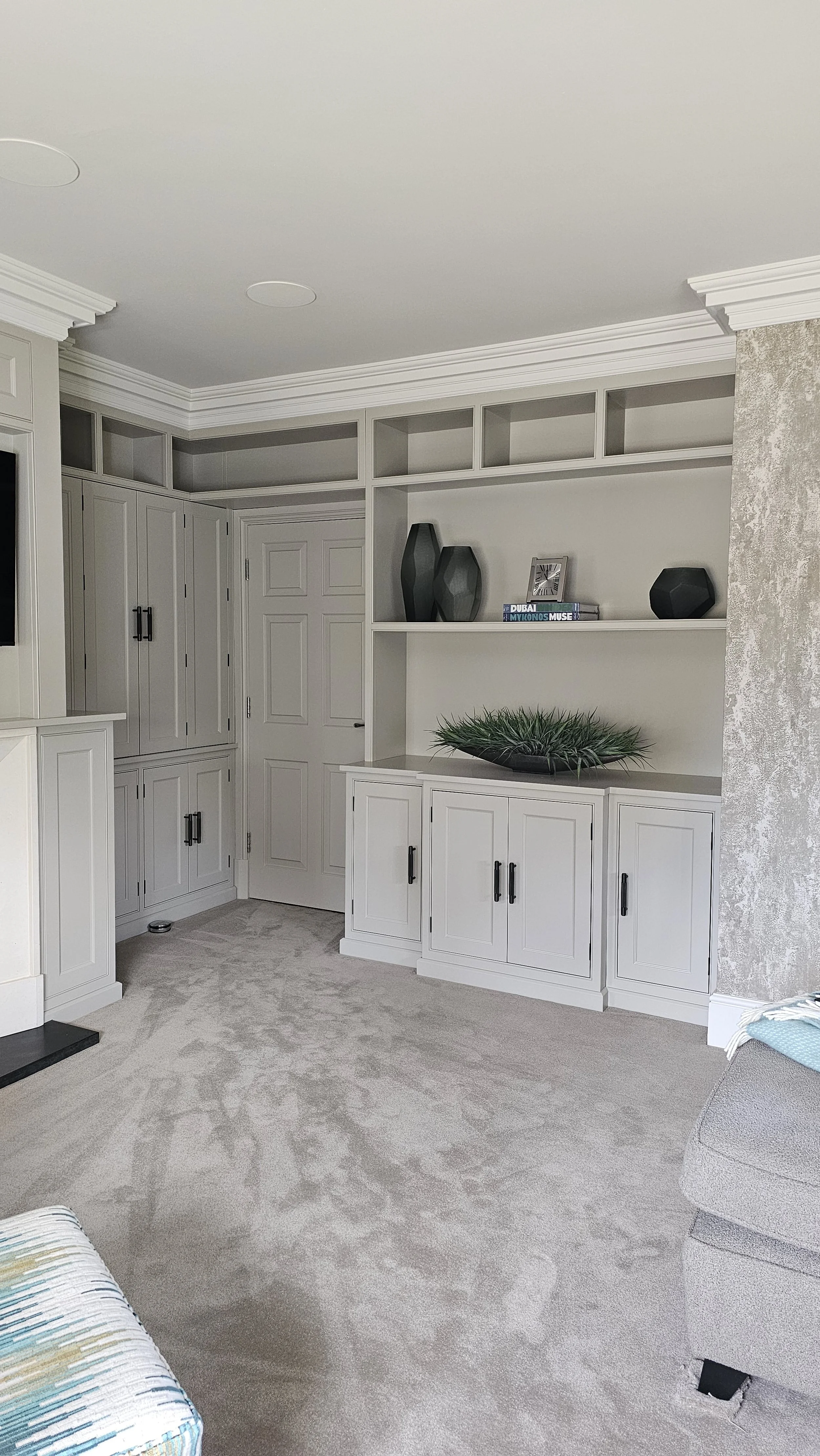 An interior living room with white built-in cabinets and a shelf above, decorated with black vases, a picture, travel books, and a large green plant on the cabinet. The room has beige carpet, a gray couch partially visible on the right, and textured 