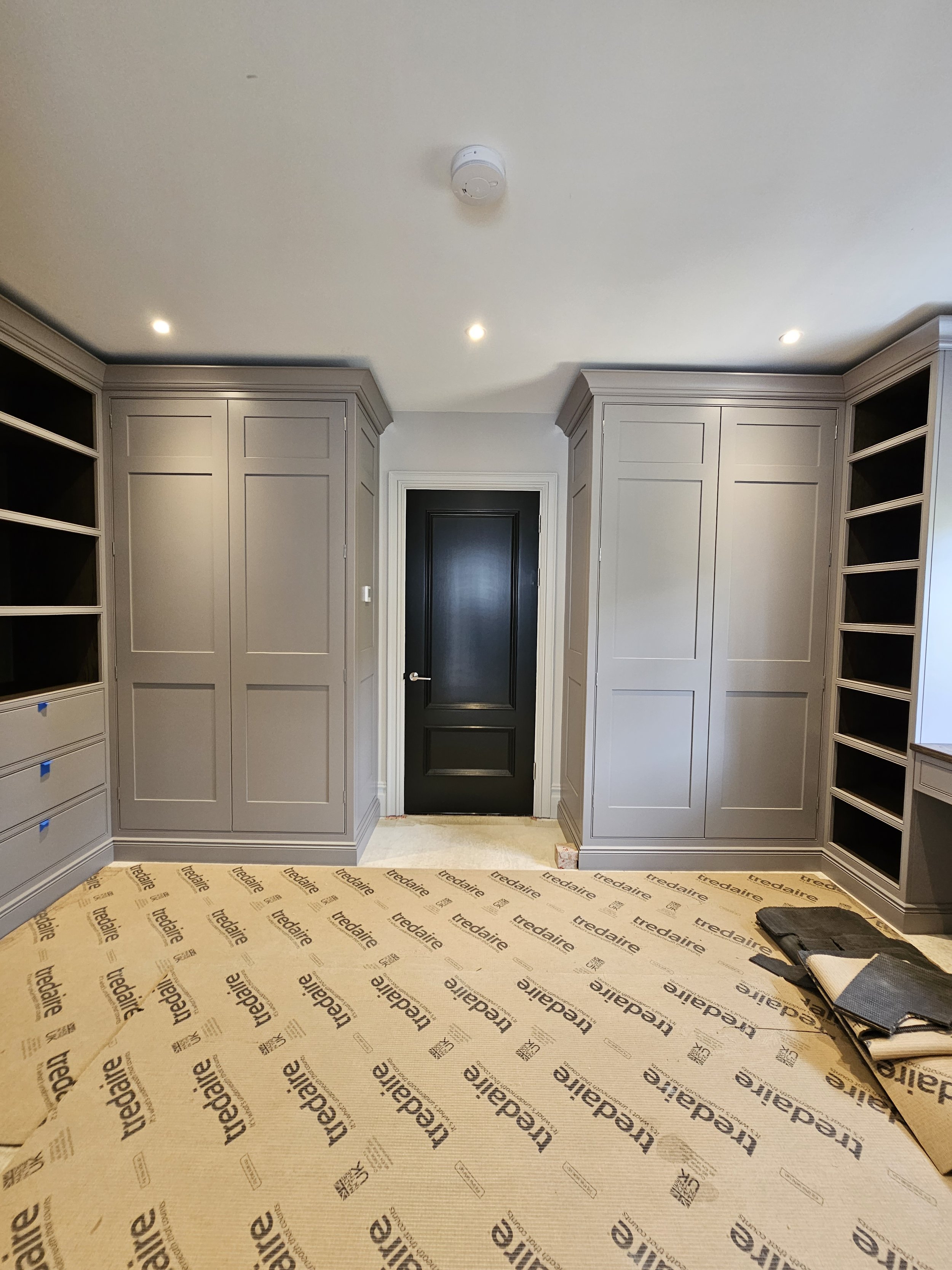 Empty room with custom gray cabinets on all sides, a black door in the center, and a stack of flooring materials on the right, indicating ongoing construction or renovation.
