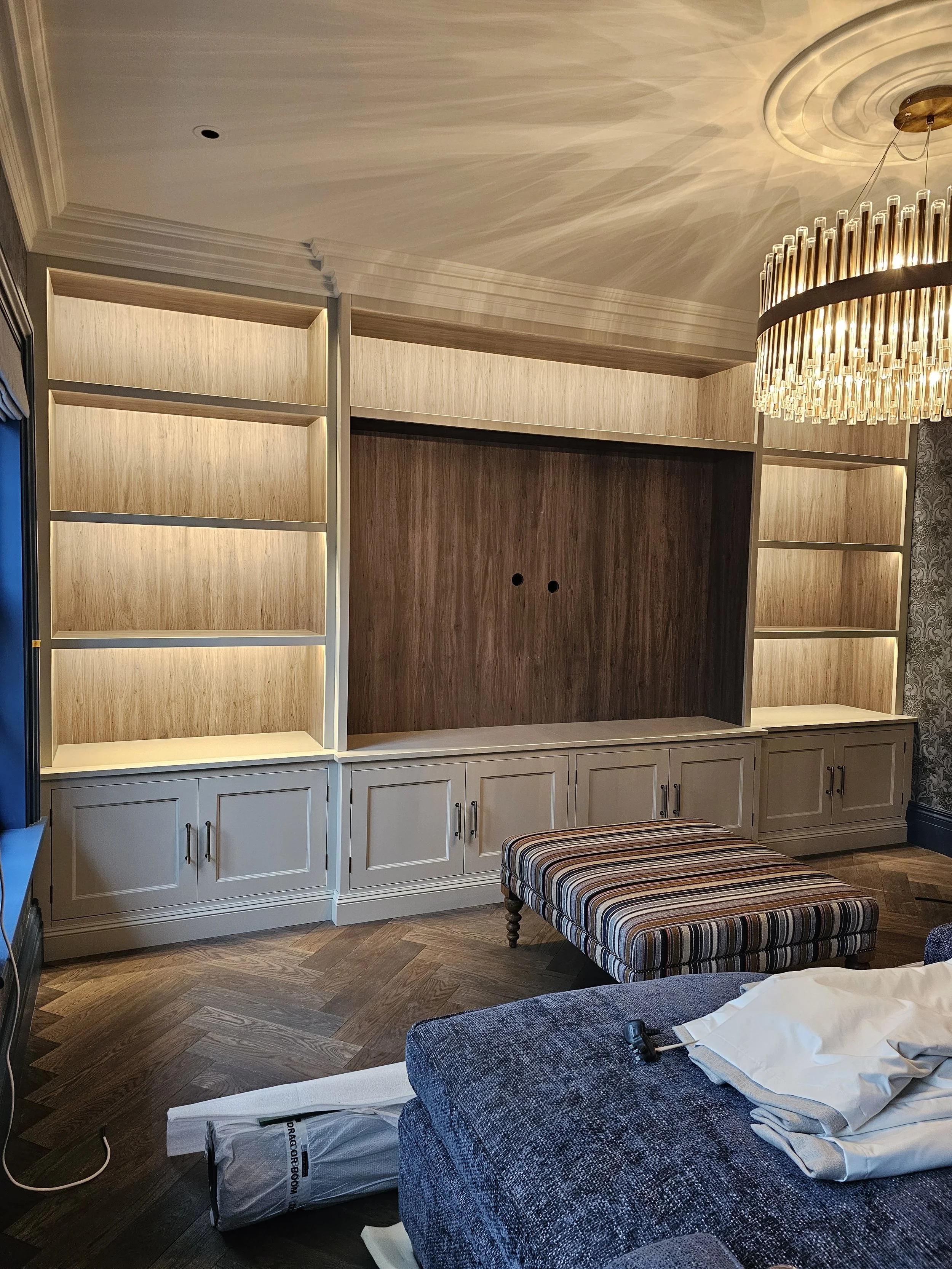 Unfinished built-in entertainment center with open shelves and cabinets below in a living room, with a striped ottoman and a blue upholstered sofa.
