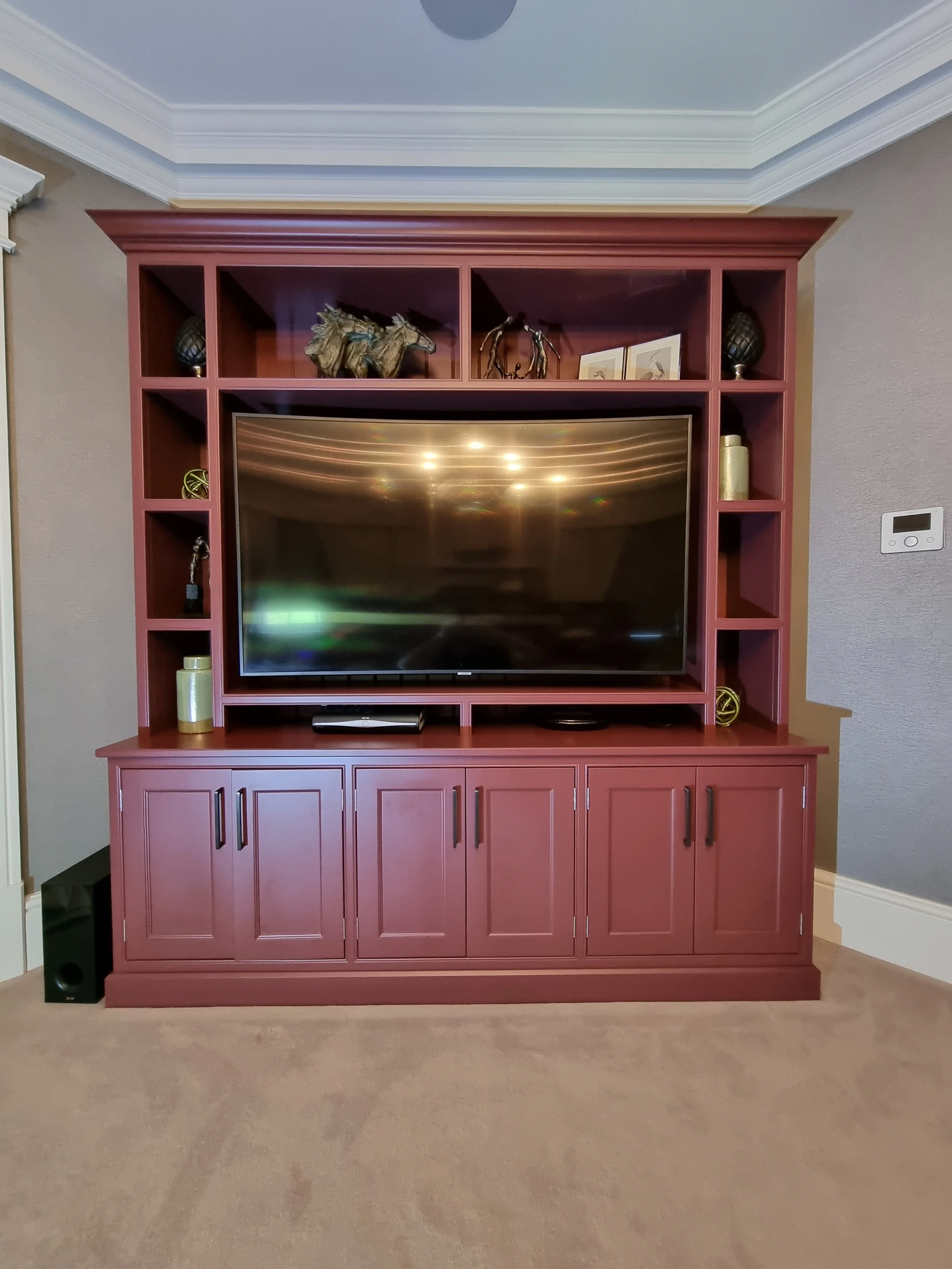 A maroon wooden entertainment center with shelves, housing a large flat-screen TV, decorative items, and a sound speaker on the floor to the left.