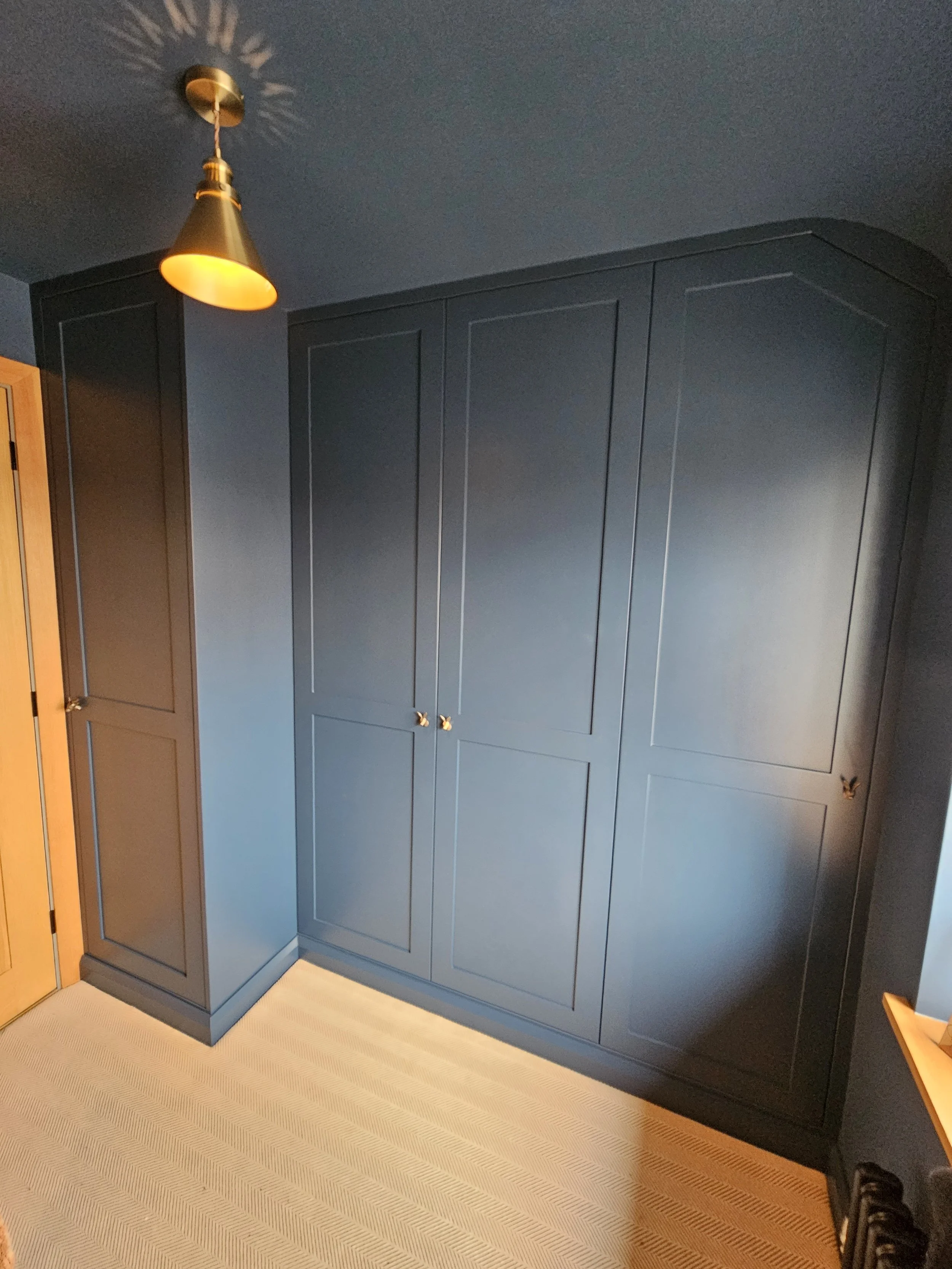 A dark blue wardrobe with four doors and brass knobs in a room with beige carpet and a wooden door on the left.