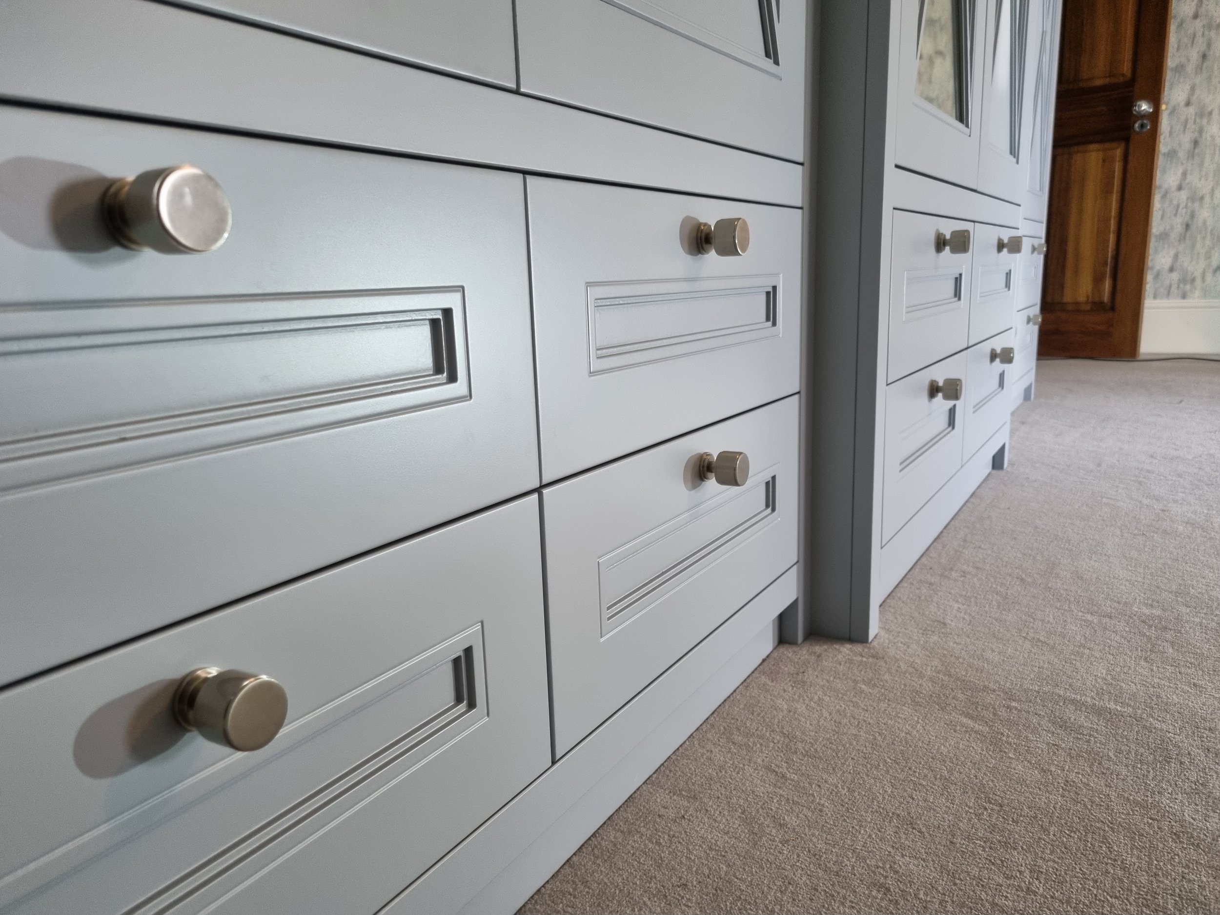 A close-up of a series of light gray cabinets with round metallic knobs, positioned on a beige carpeted floor in an interior space.