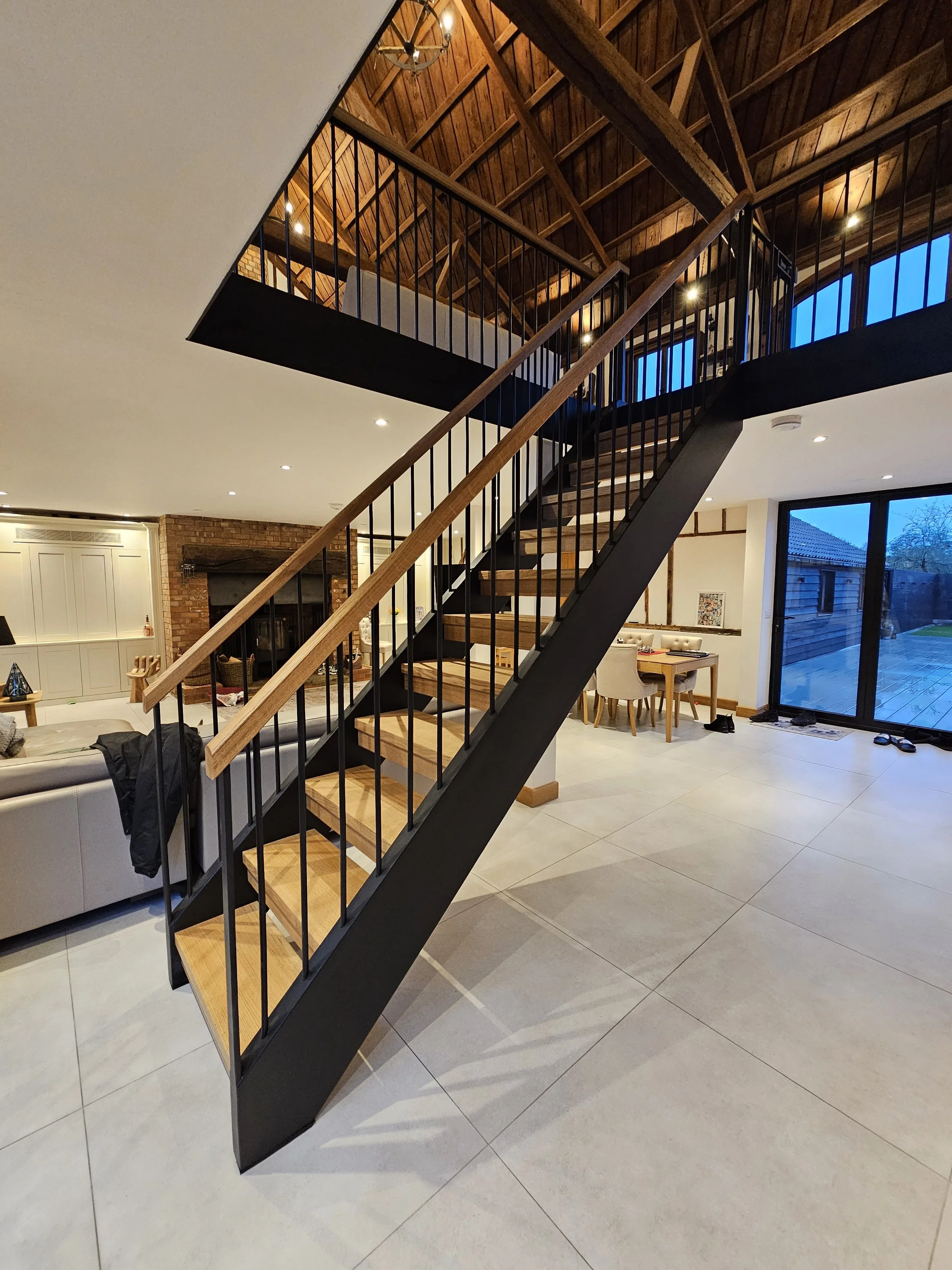 Interior view of a modern house with a wooden staircase leading to an upstairs balcony, featuring wooden flooring and black railings, with a living area, dining table, and sliding glass doors visible.