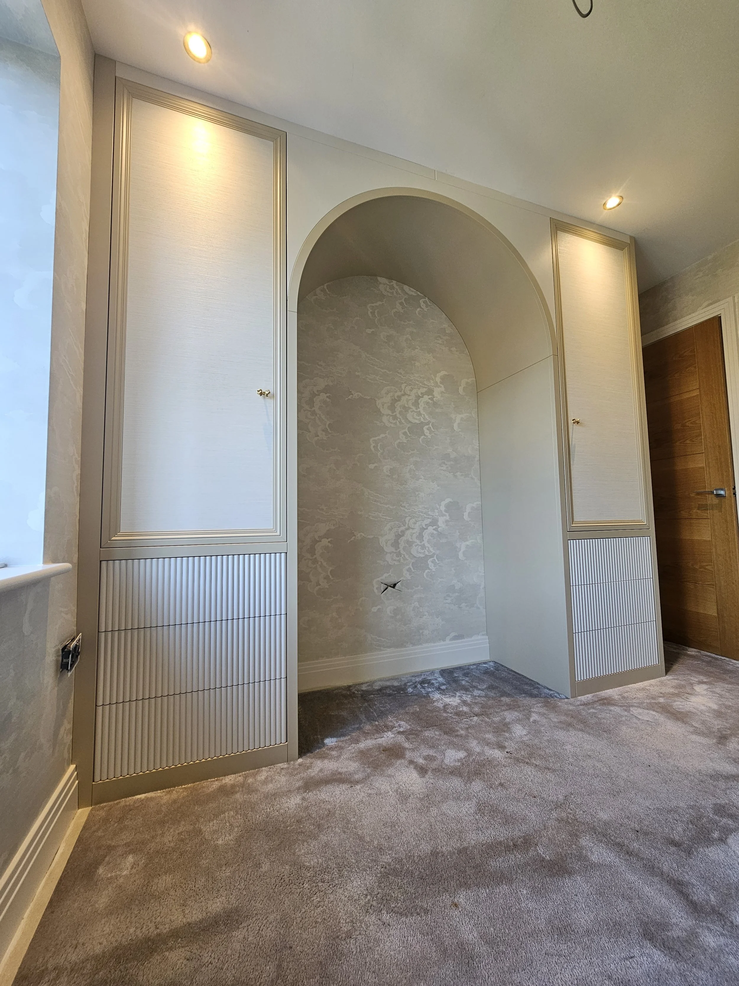 Empty room with built-in cabinets and an archway, partially decorated wall, carpeted floor, near a window, with ceiling spotlights and a wooden door.