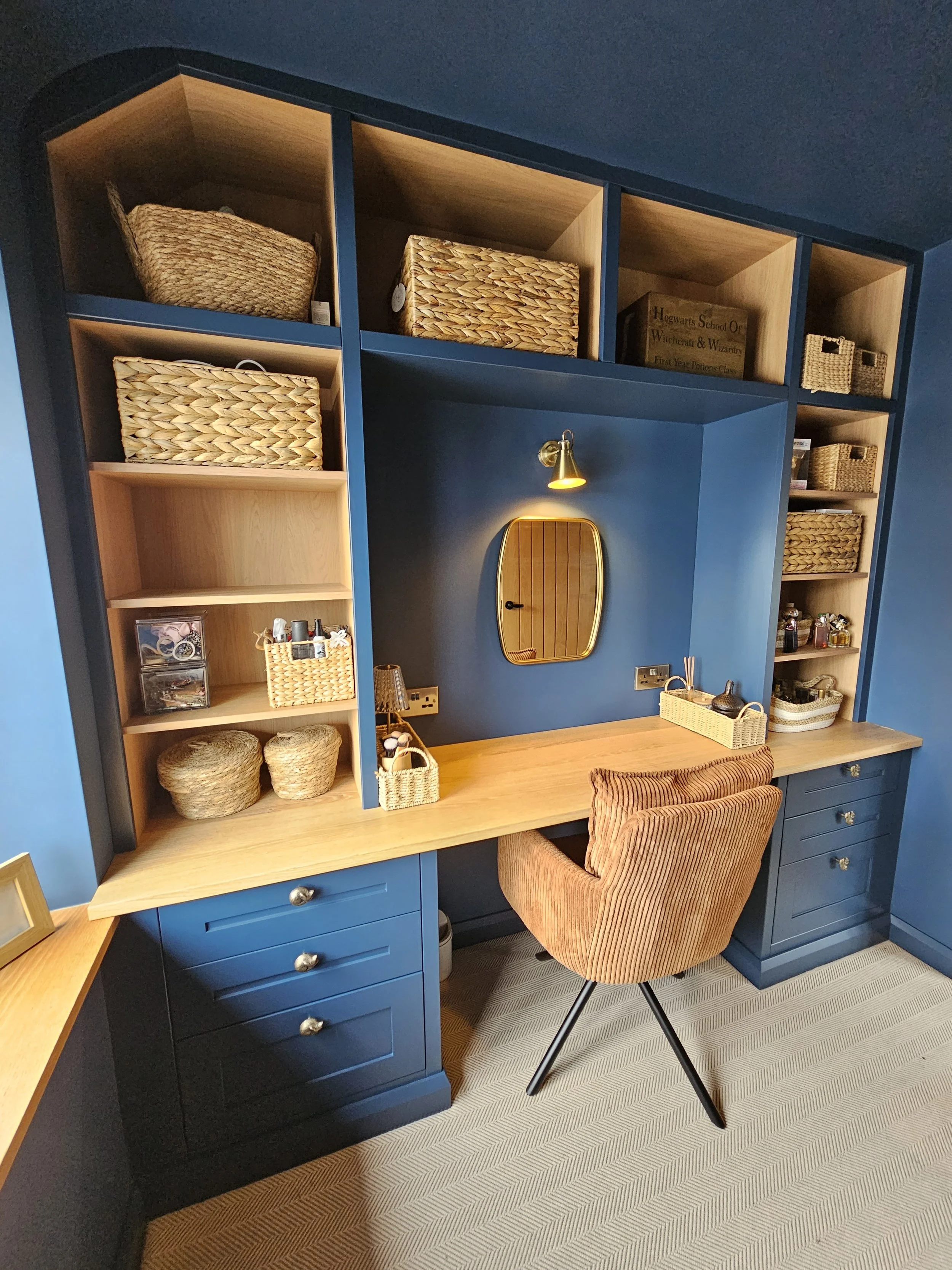 A corner of a home office with a blue wall and built-in shelves. The shelves contain wicker baskets and decor. There is a wooden desk with a brown upholstered chair, a mirror on the wall, and a small light above it.