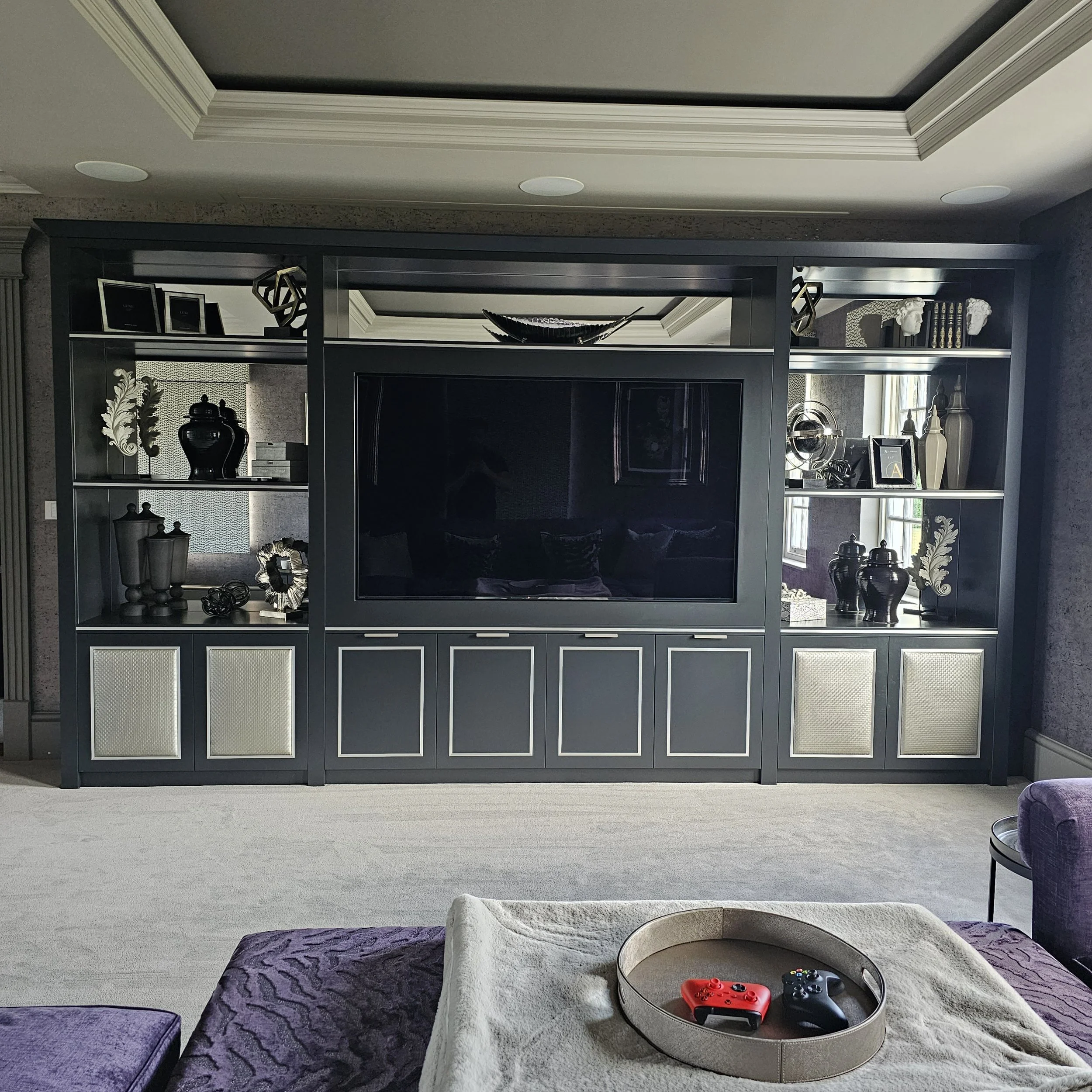 Living room with a black entertainment center, decorated with vases, framed photos, and decorative objects, with a flat-screen TV, purple furniture, and a tray holding video game controllers.