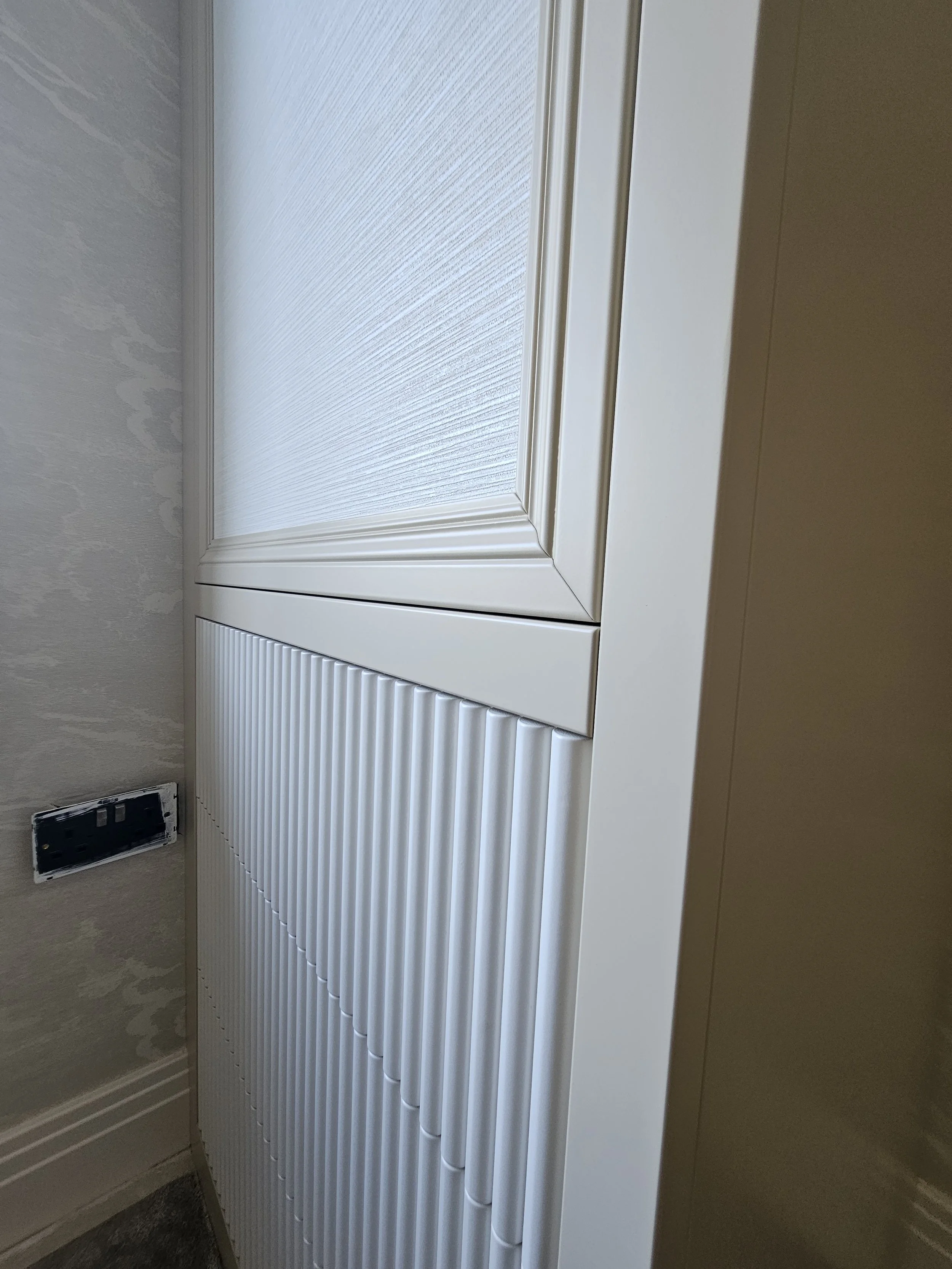 Close-up image of a white cabinet door with textured panels and beveled edges next to an electrical outlet on a beige wall.