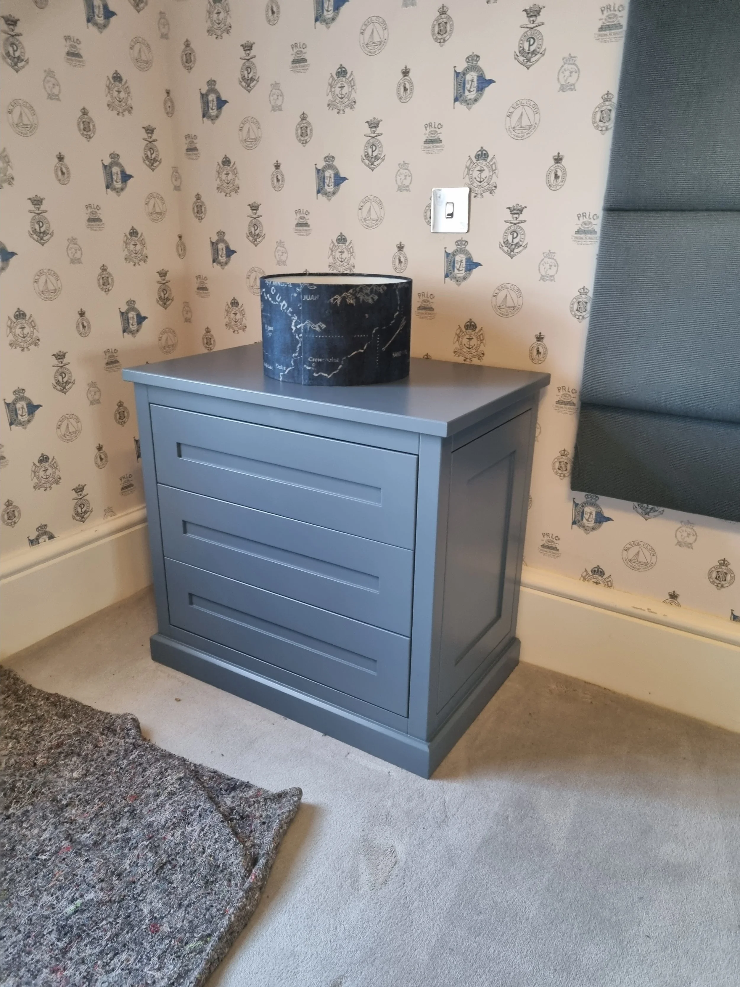 A gray nightstand with four drawers is placed next to a wall decorated with wallpaper featuring a blueprint and crest design. There is a black marble container on top of the nightstand. A power outlet is visible on the wall.