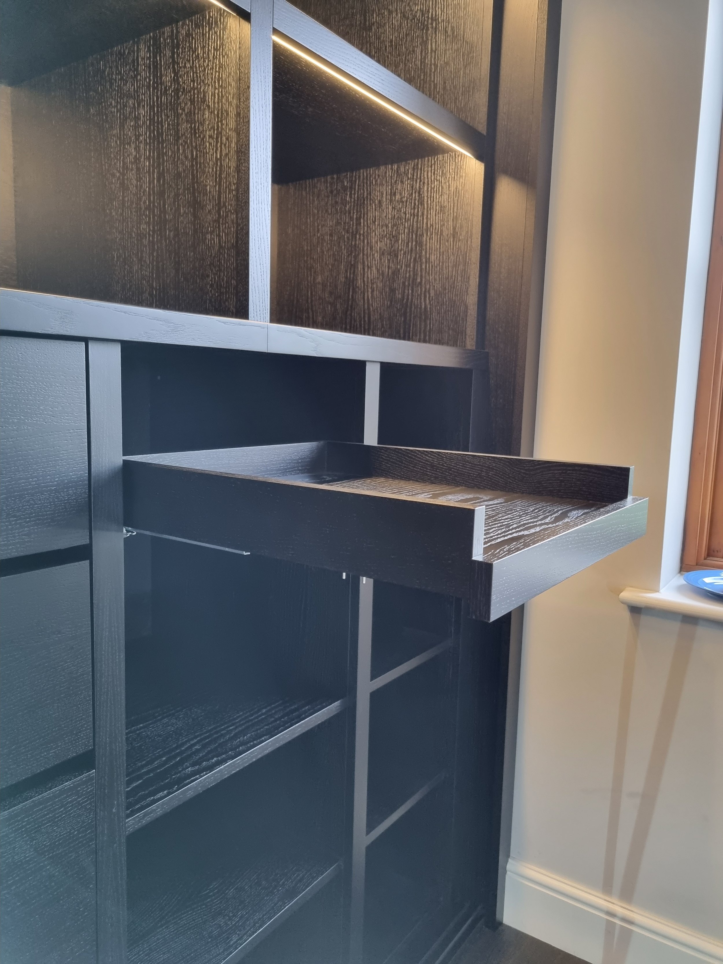 A black wooden shelf unit with open shelves, partially pulled out, located near a window with a blue plate on the windowsill.