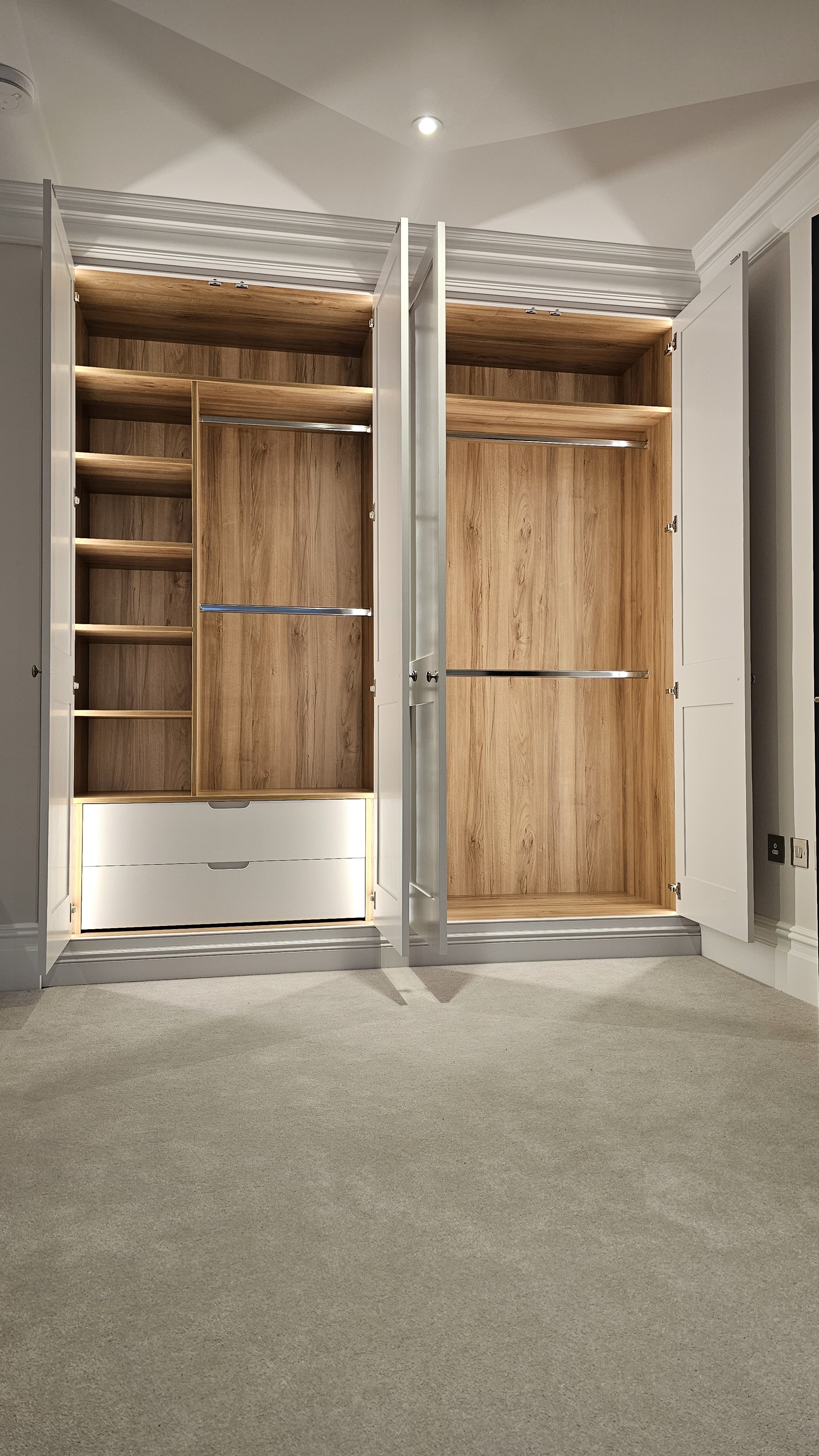 Open wardrobe closet with white doors, wooden interior, shelves, drawers, and hanging space in a room with beige carpet.