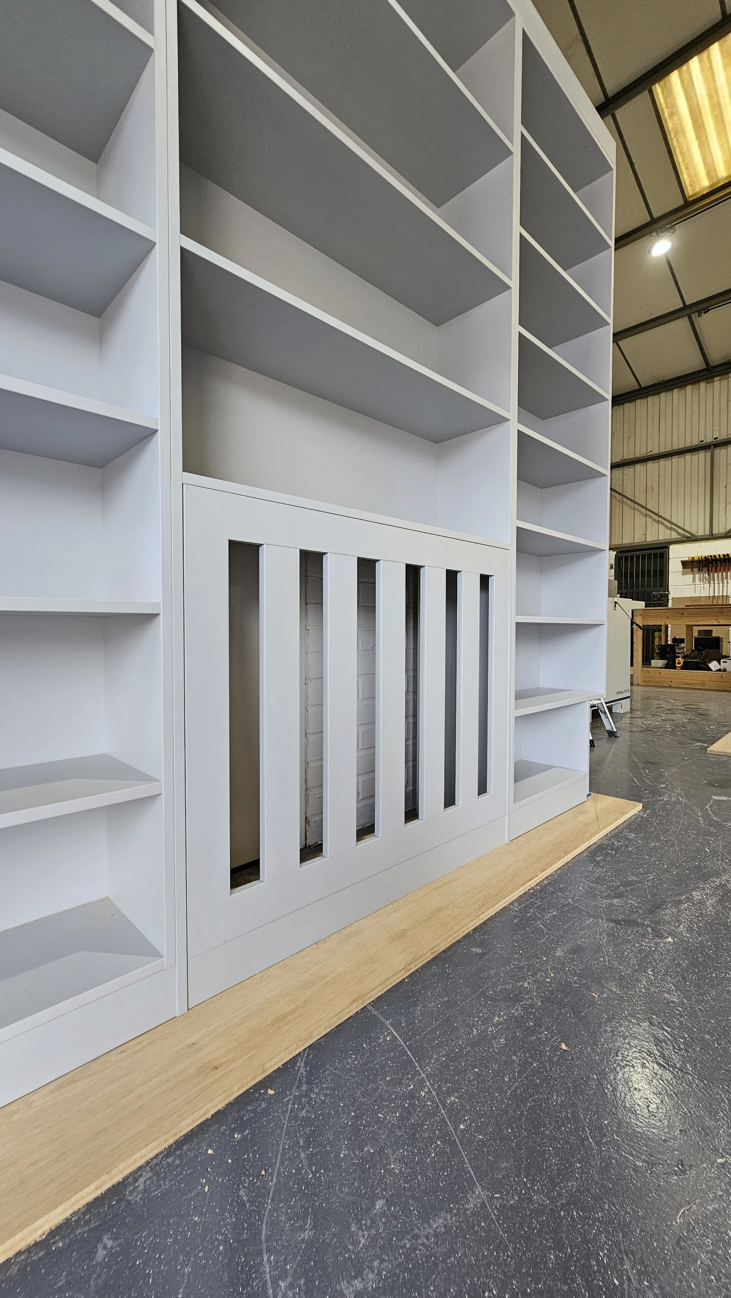 White built-in bookshelf with multiple shelves in a workshop or warehouse, with an unfinished wooden base and exposed brick wall behind.