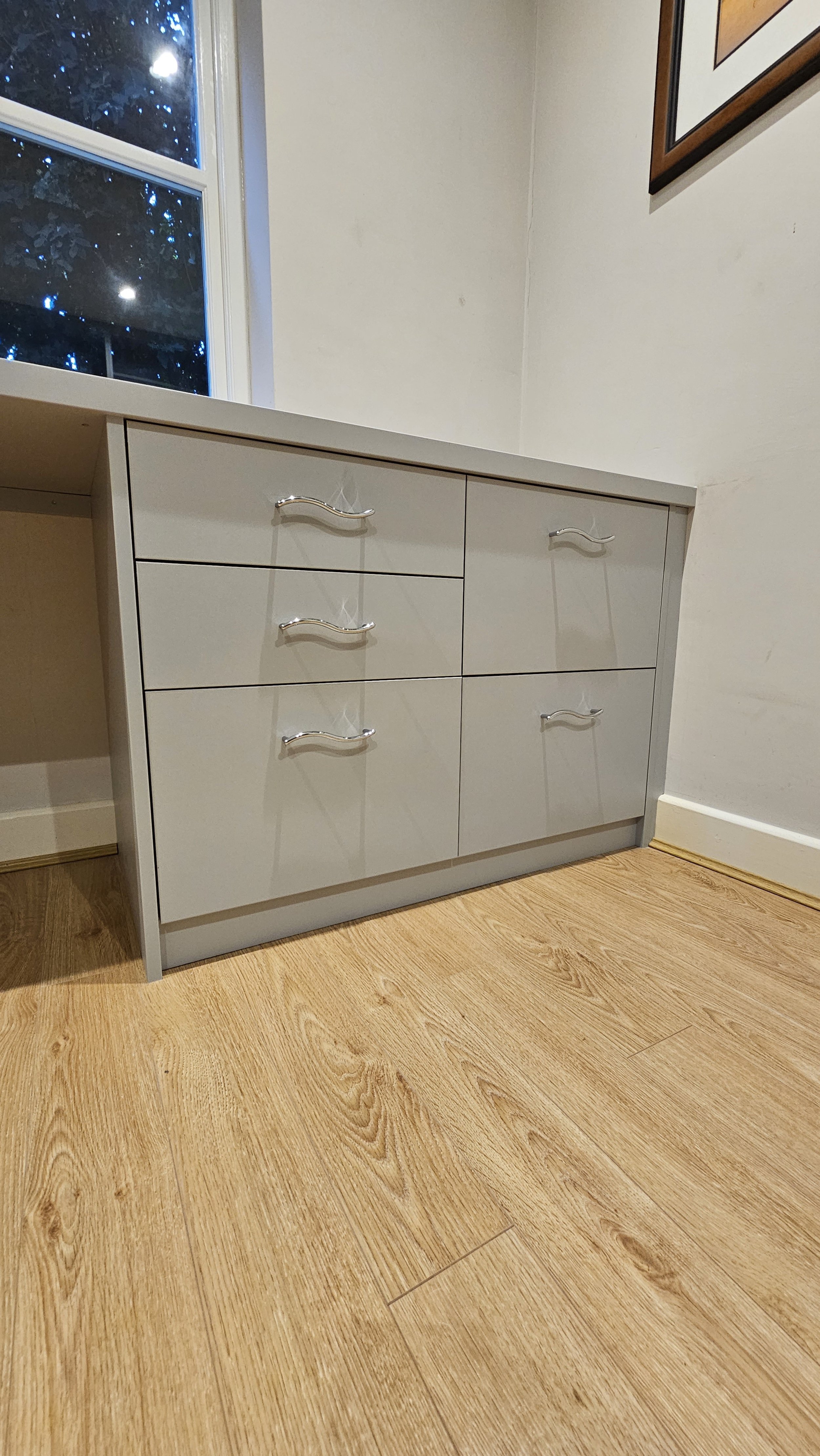Gray dresser with six drawers and curved silver handles, situated on wooden floor next to a wall with a window showing trees outside.
