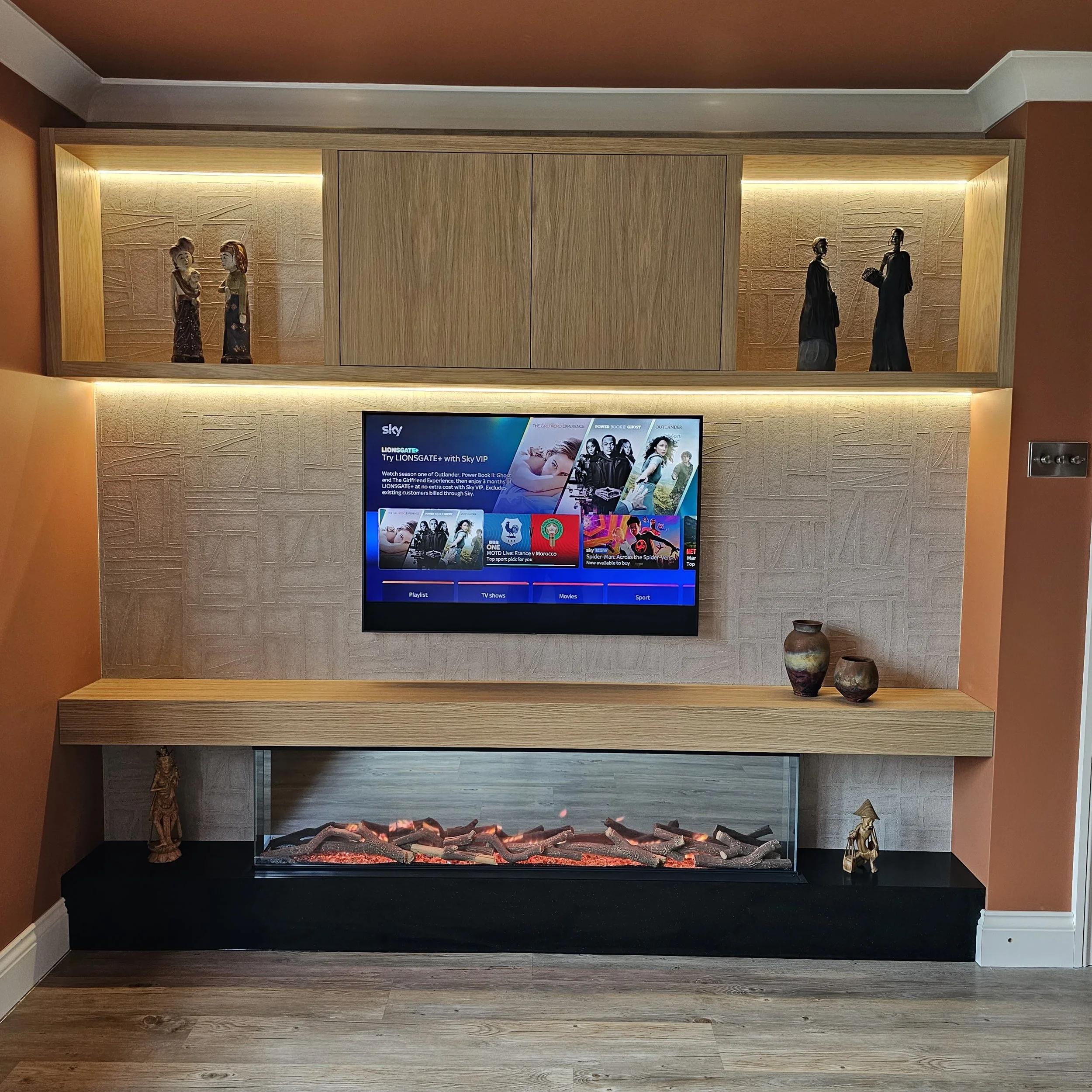 Living room wall with mounted flat-screen TV displaying a streaming service menu, surround sound speakers, wooden and decorative pottery items, and an electric fireplace with artificial logs.