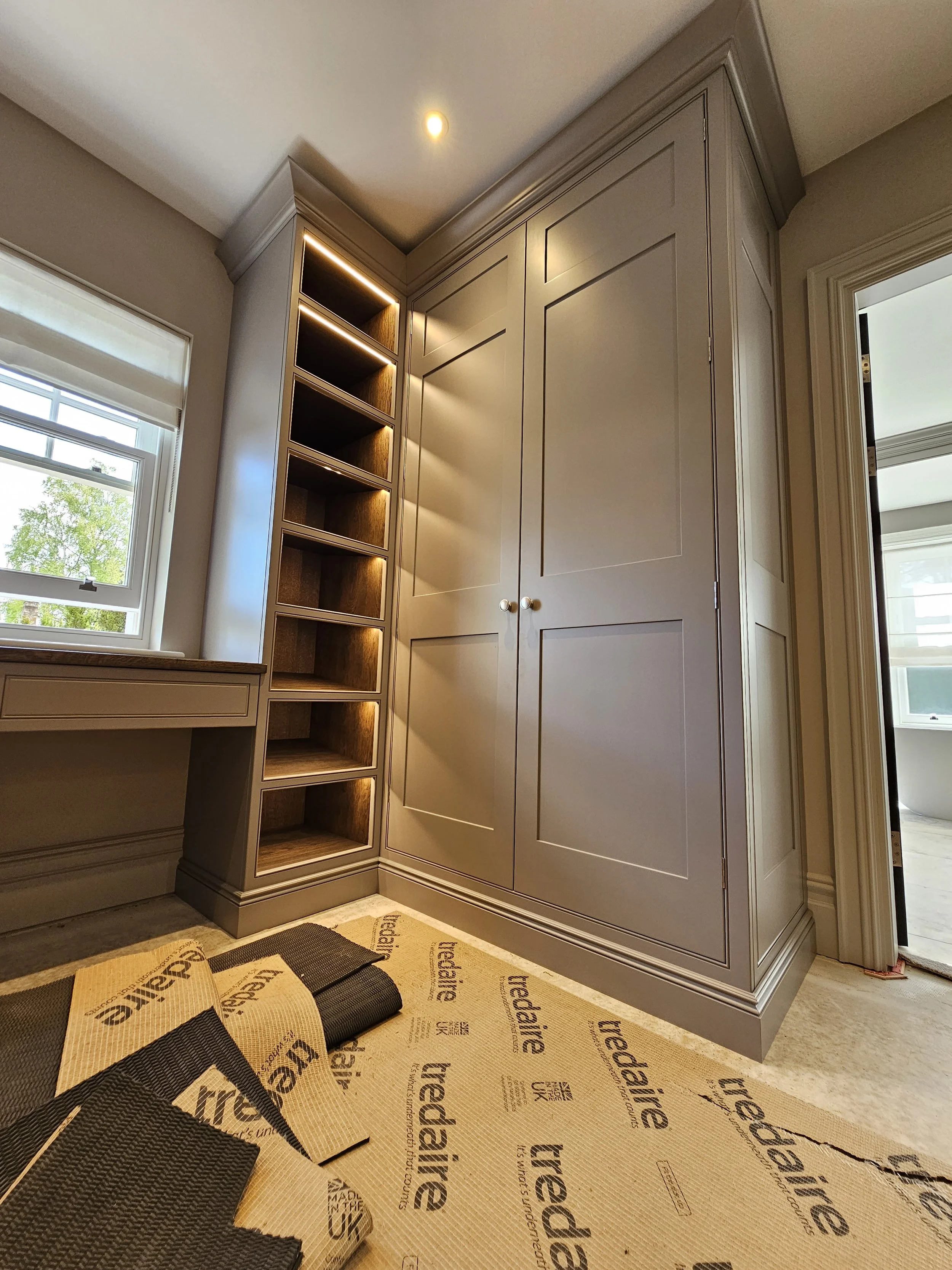 Interior of a room under construction featuring a built-in corner cabinet with closed doors and open shelves with warm lighting, a window with a view outside, and a door leading to a bathroom. Floor is covered with protective material.