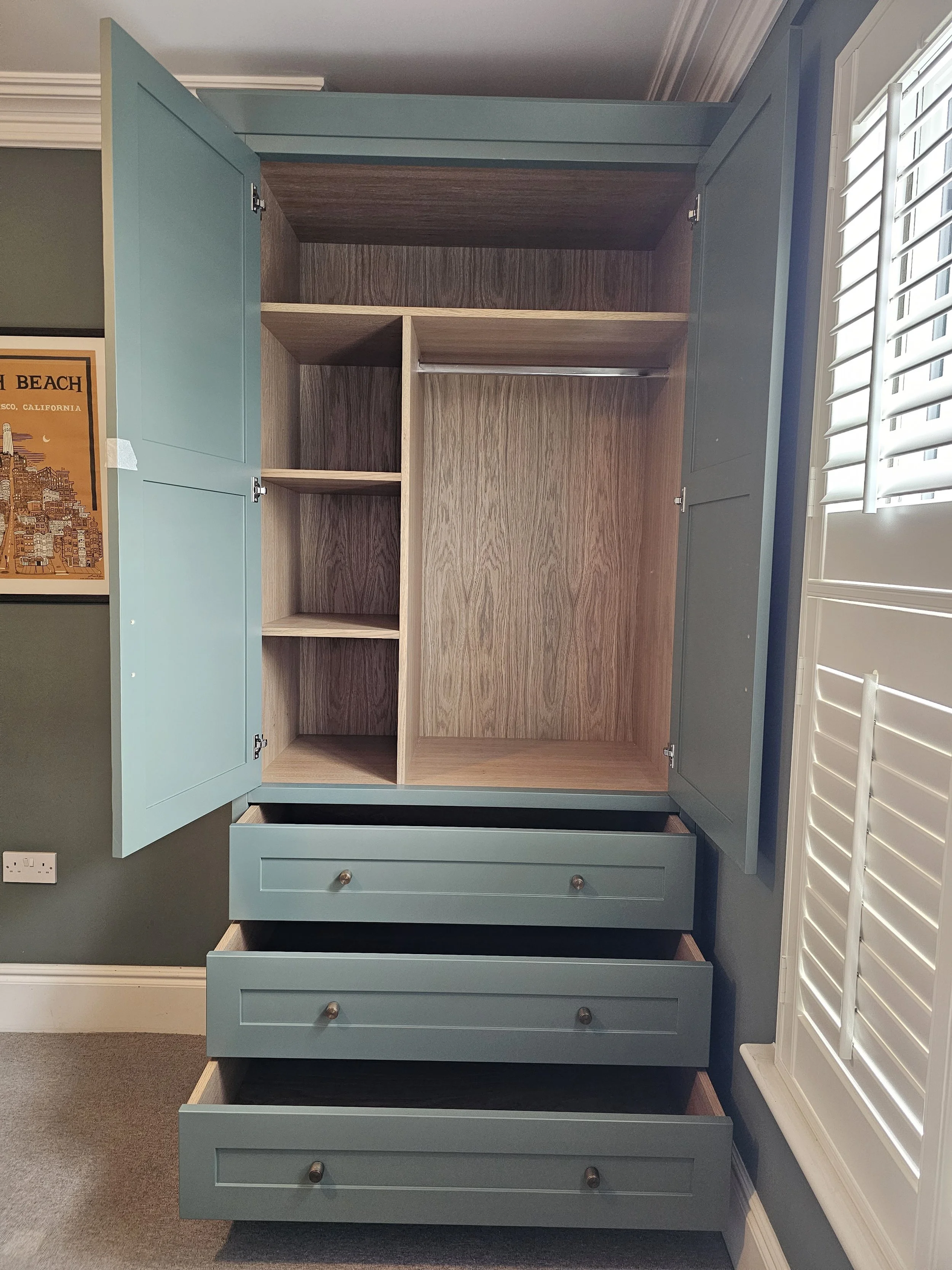 Open blue wardrobe with multiple drawers and empty shelves, located next to a window with white shutters.