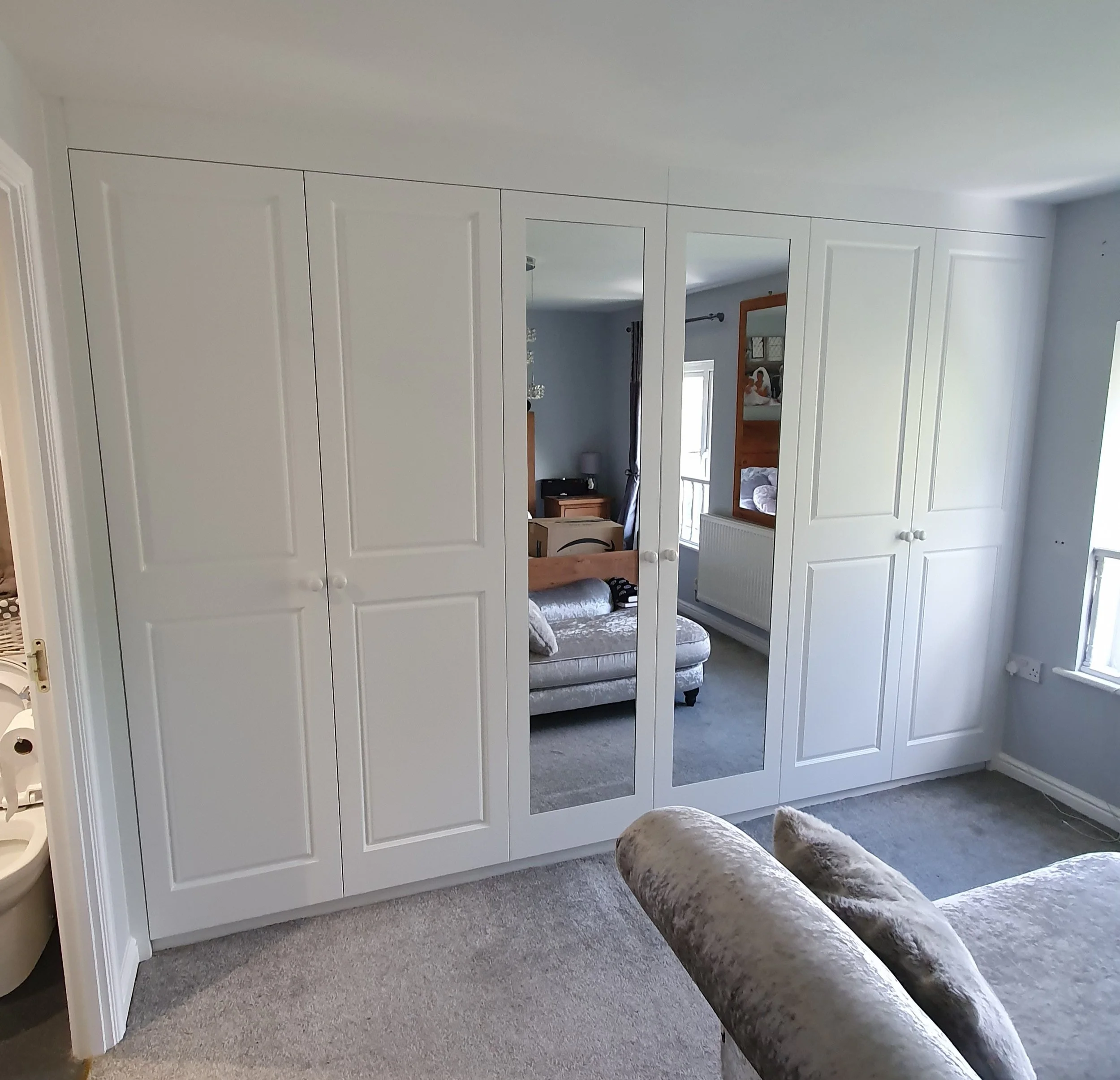 A white wardrobe with four doors, including two mirror doors, in a bedroom with a beige carpet and a light gray wall. Part of a plush beige armchair is visible in the foreground.