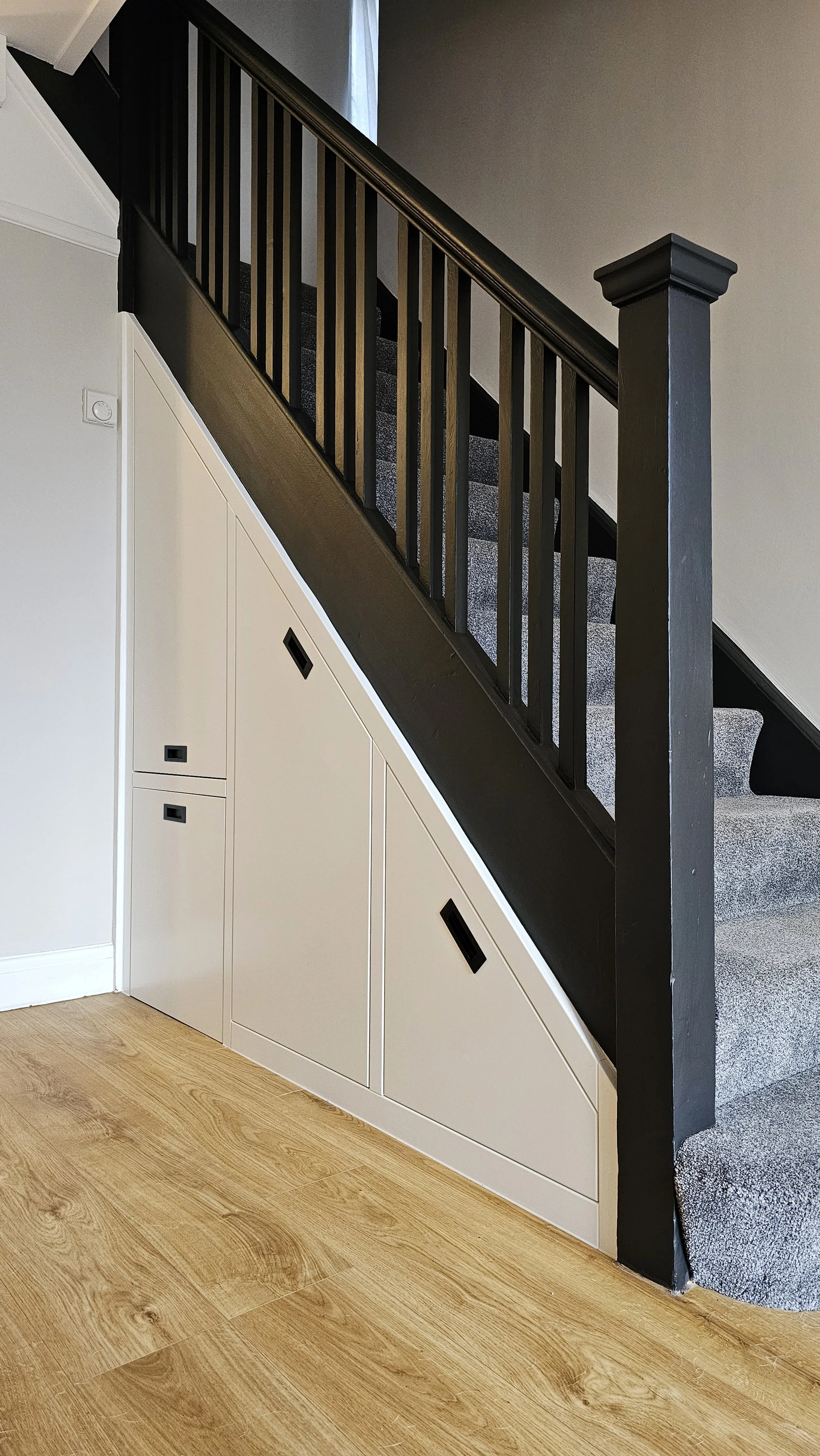 Interior view of a staircase with black wooden railing and gray carpeted steps. White built-in storage cabinets with black handles are under the stairs, and a light wood floor is visible.