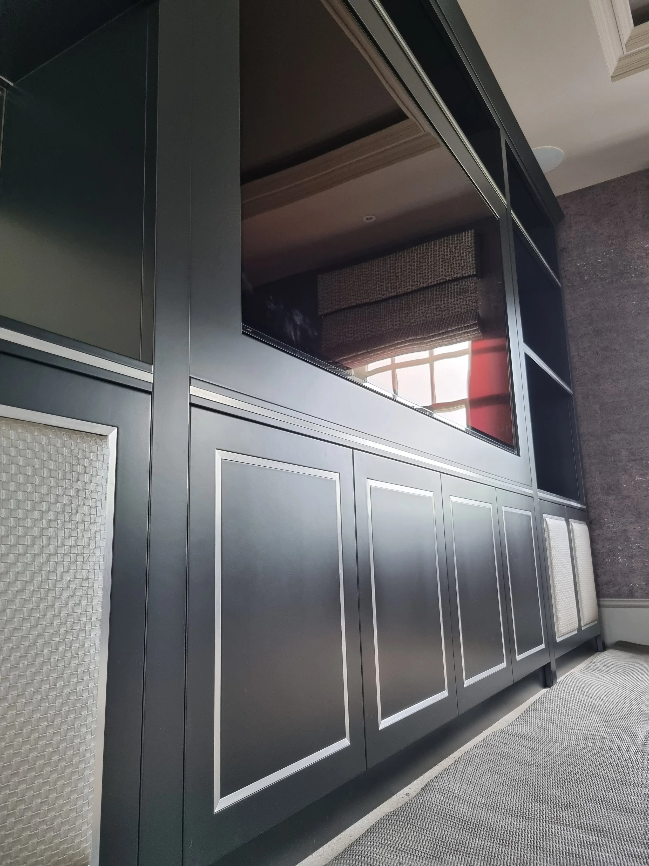 Close-up view of a black built-in cabinet with rectangular panels and metallic trim, positioned along a textured wall and under a glass window, with part of a carpeted floor in the foreground.