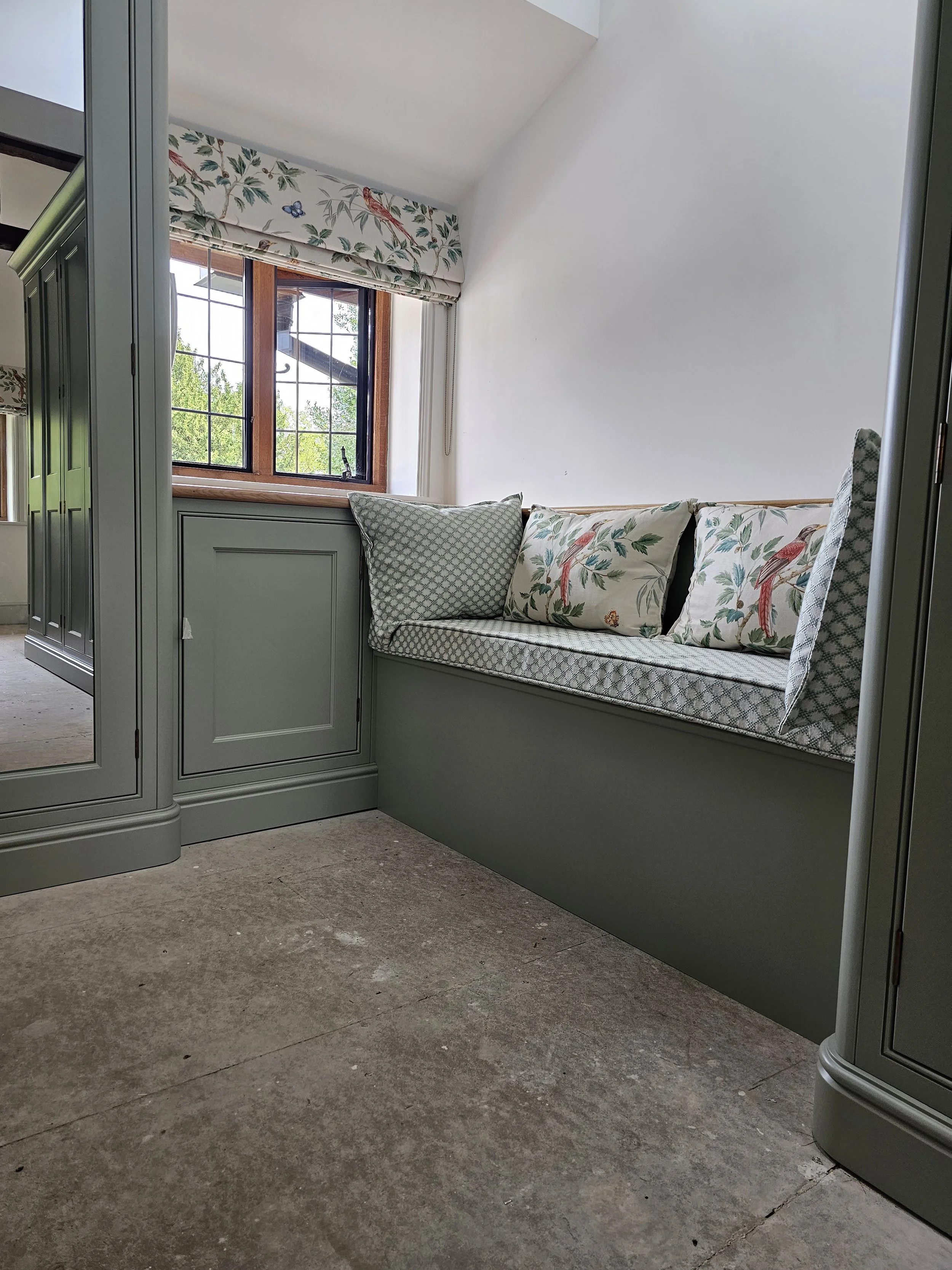 A cozy window seat with cushions and pillows, positioned beside a window with a floral valance, in a room with light-colored walls and a stone floor.