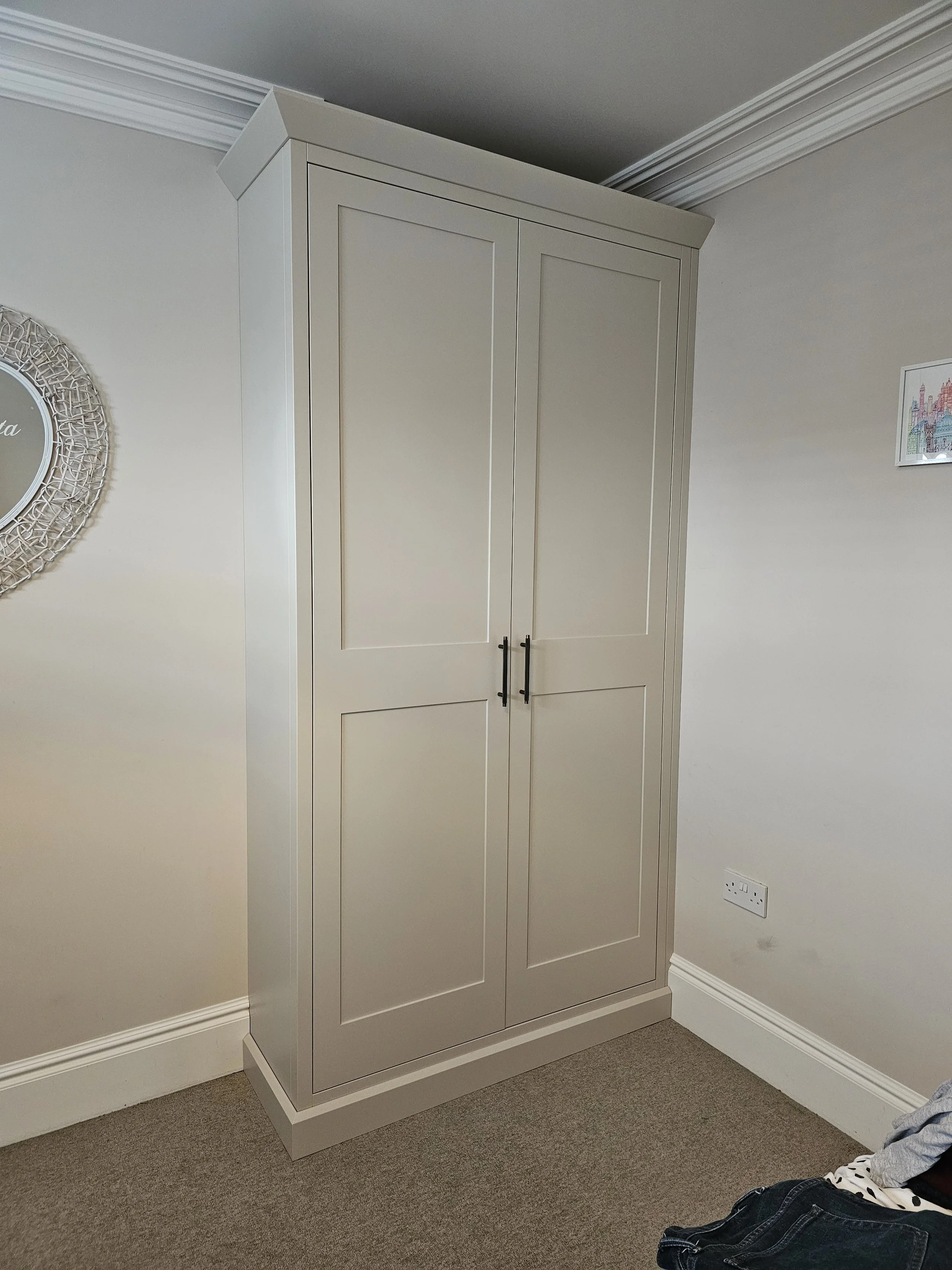 A white wardrobe with two doors and black handles standing in a corner of a room with cream-colored walls, beige carpet, crown molding, and a double electrical outlet.