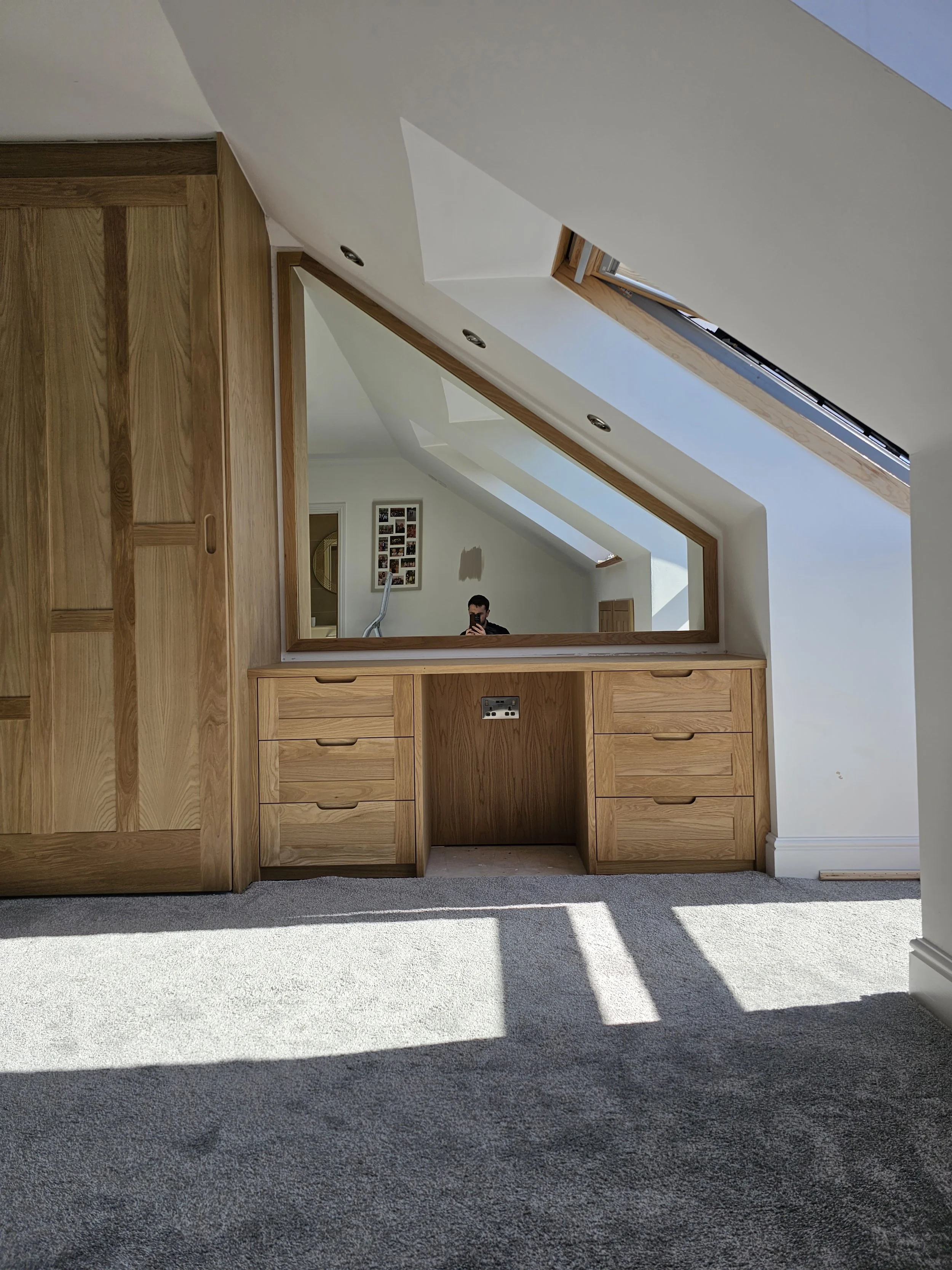 Custom wooden desk in an attic with sloped ceilings and skylights, reflected in a large mirror on the wall.