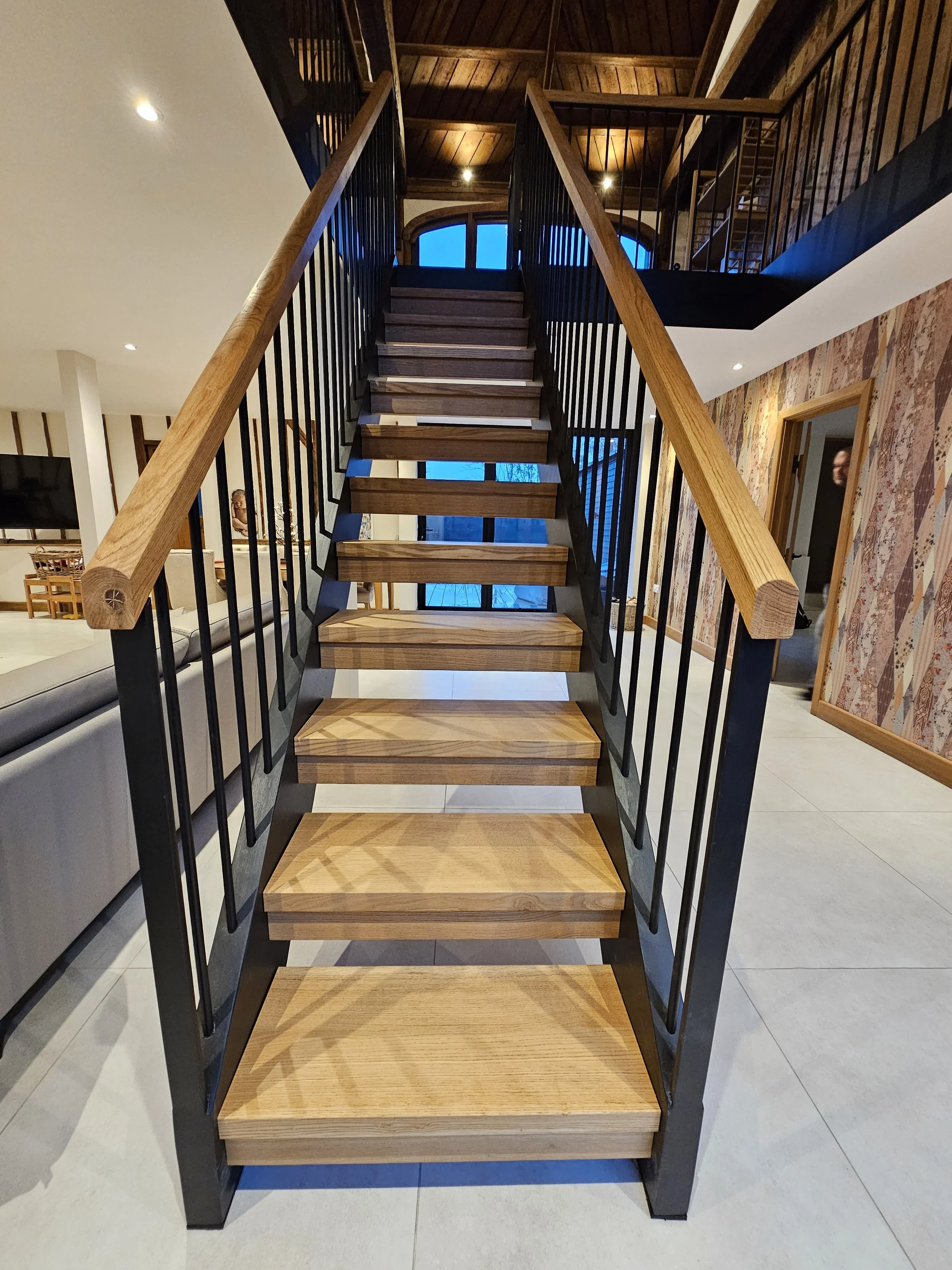 Indoor staircase made of wood and black metal with a wooden handrail, leading up to a second floor in a modern home with wood-paneled ceiling and decorative wallpaper on the walls.
