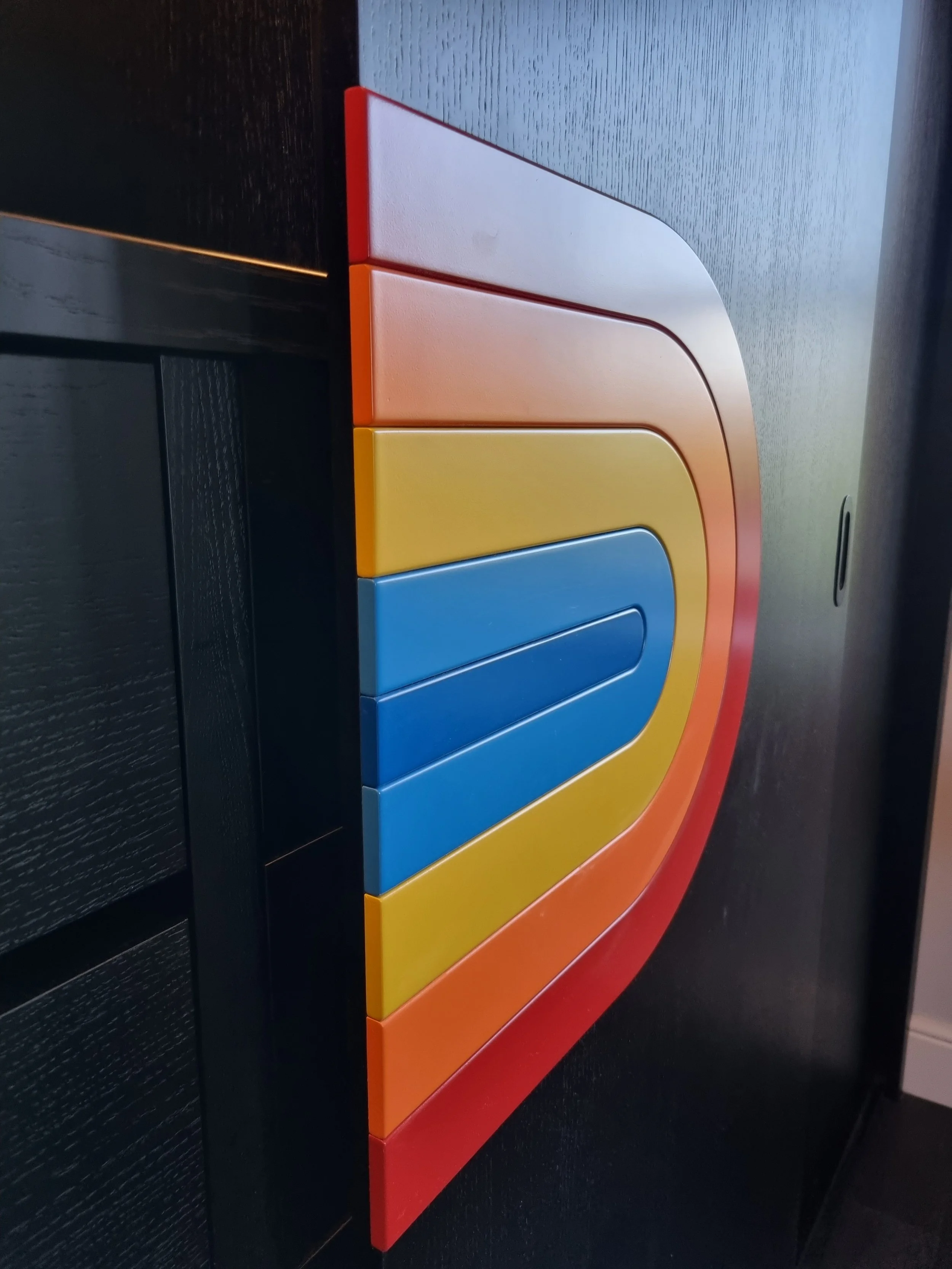 Colorful children’s wall-mounted coat hooks in red, orange, yellow, and blue, attached to a black wall.