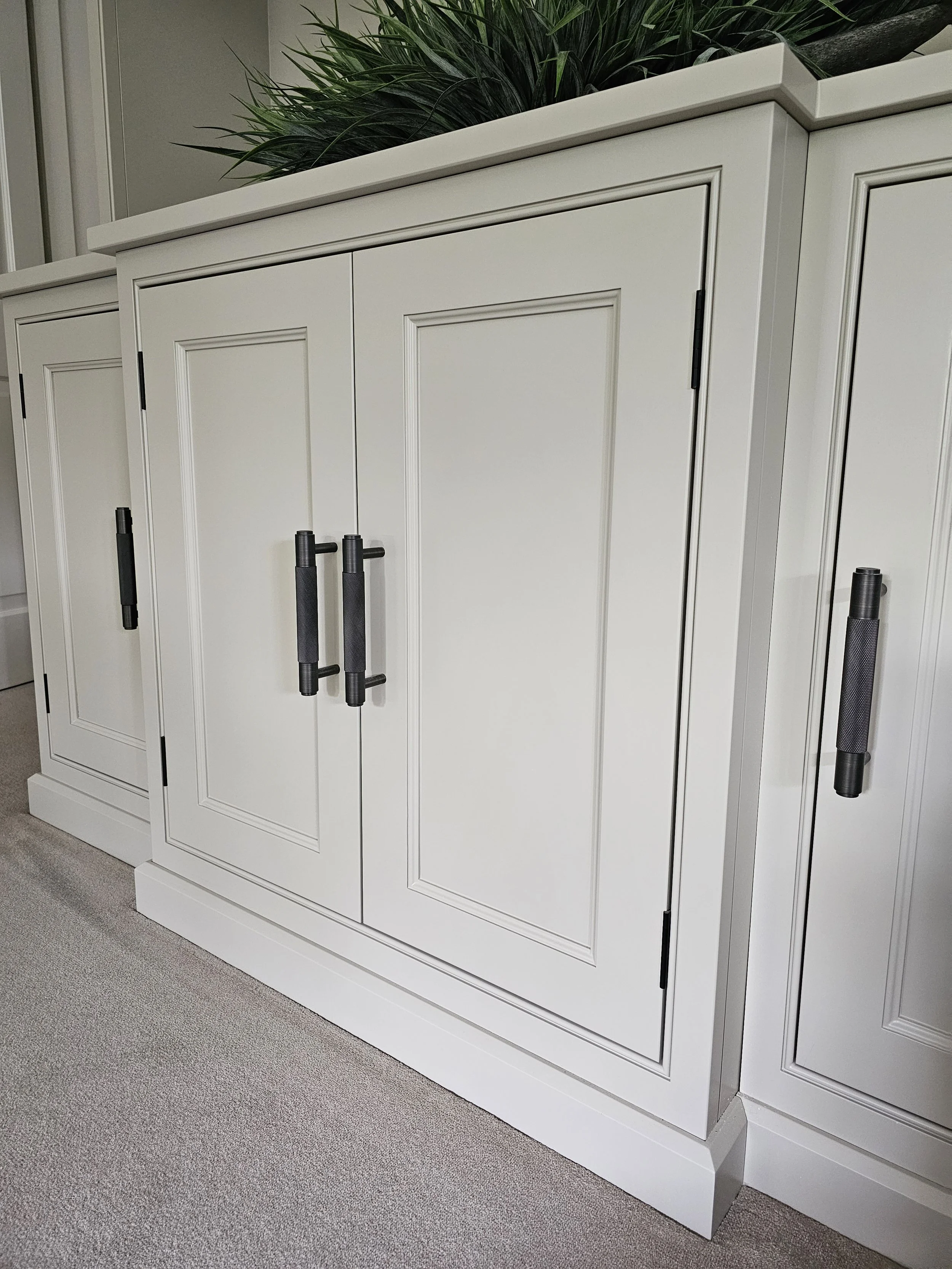 White cabinet with paneled doors and black hardware handles, topped with green plants.