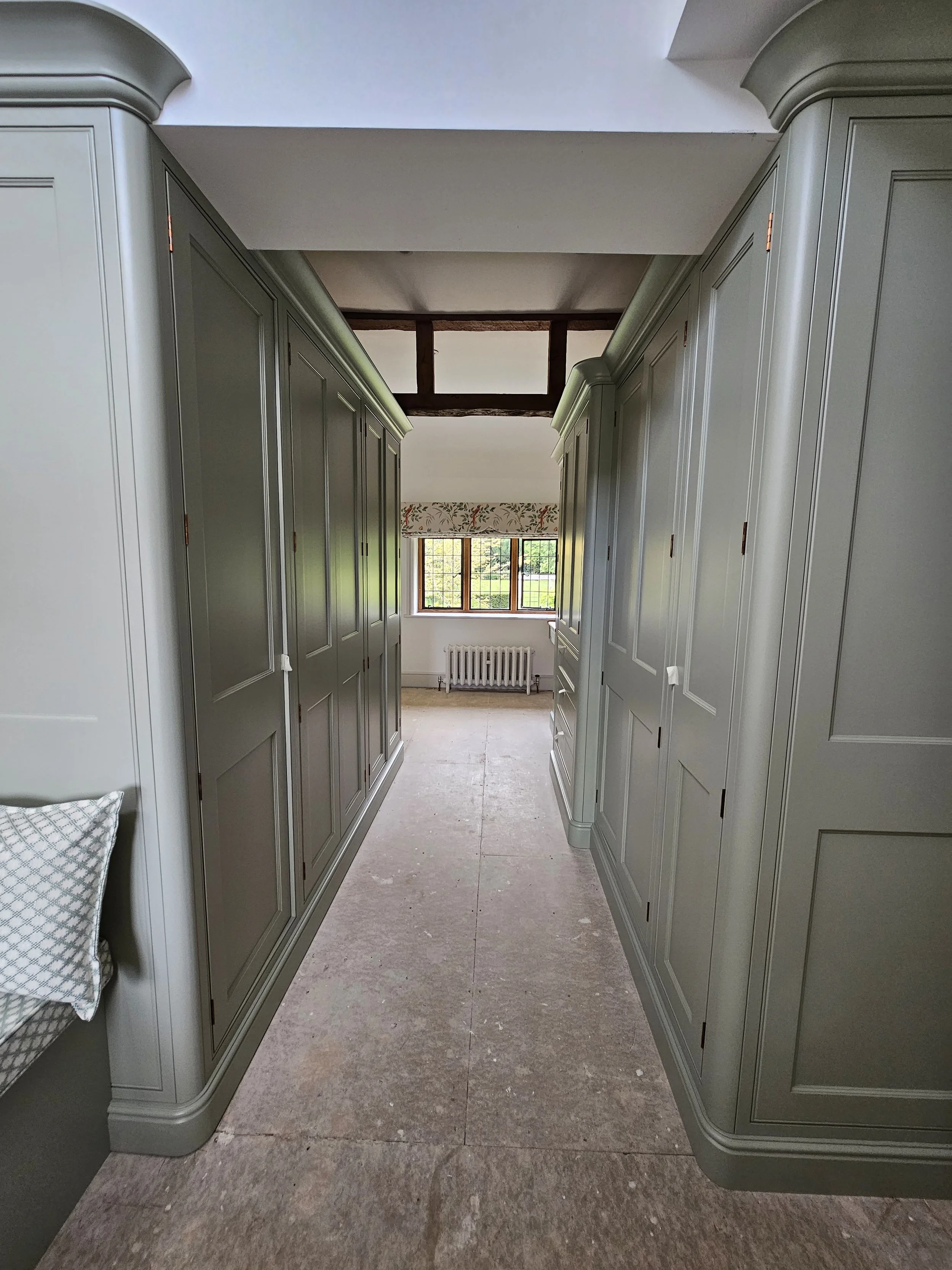 A hallway with built-in, light green cabinets on both sides, leading to a room with a window, radiator, and floral valance.
