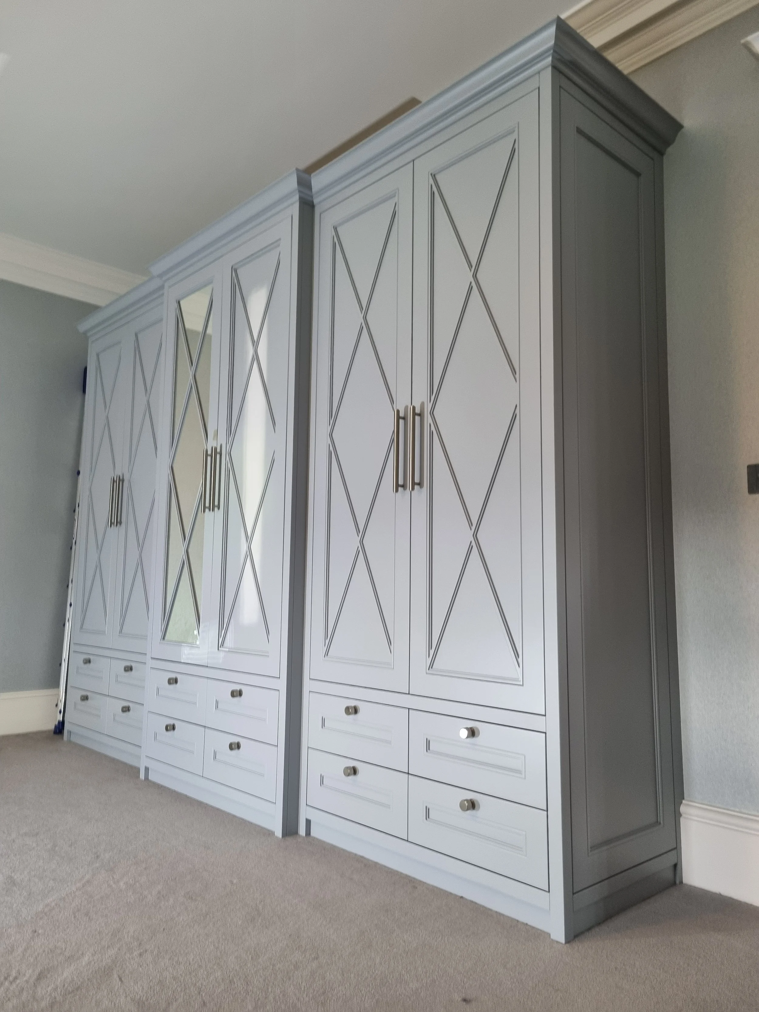 White and gray wardrobe with mirrored doors, decorative paneling, and multiple drawers, positioned against a light wall with carpeted floor.