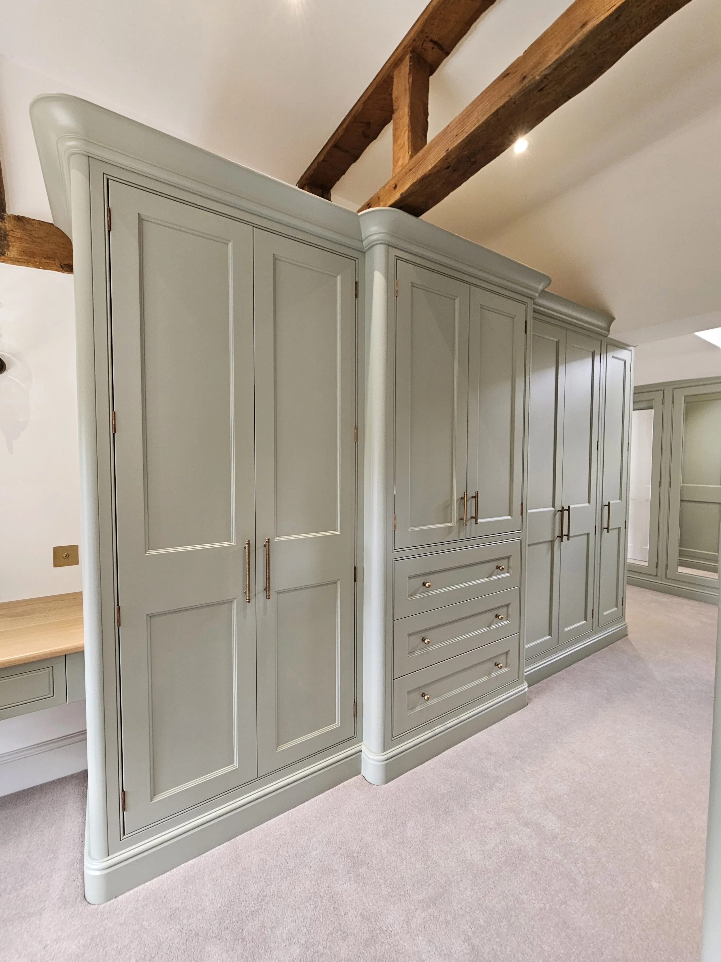 A large custom-built light gray wardrobe with gold handles, situated in a room with wood ceiling beams and a beige carpet.