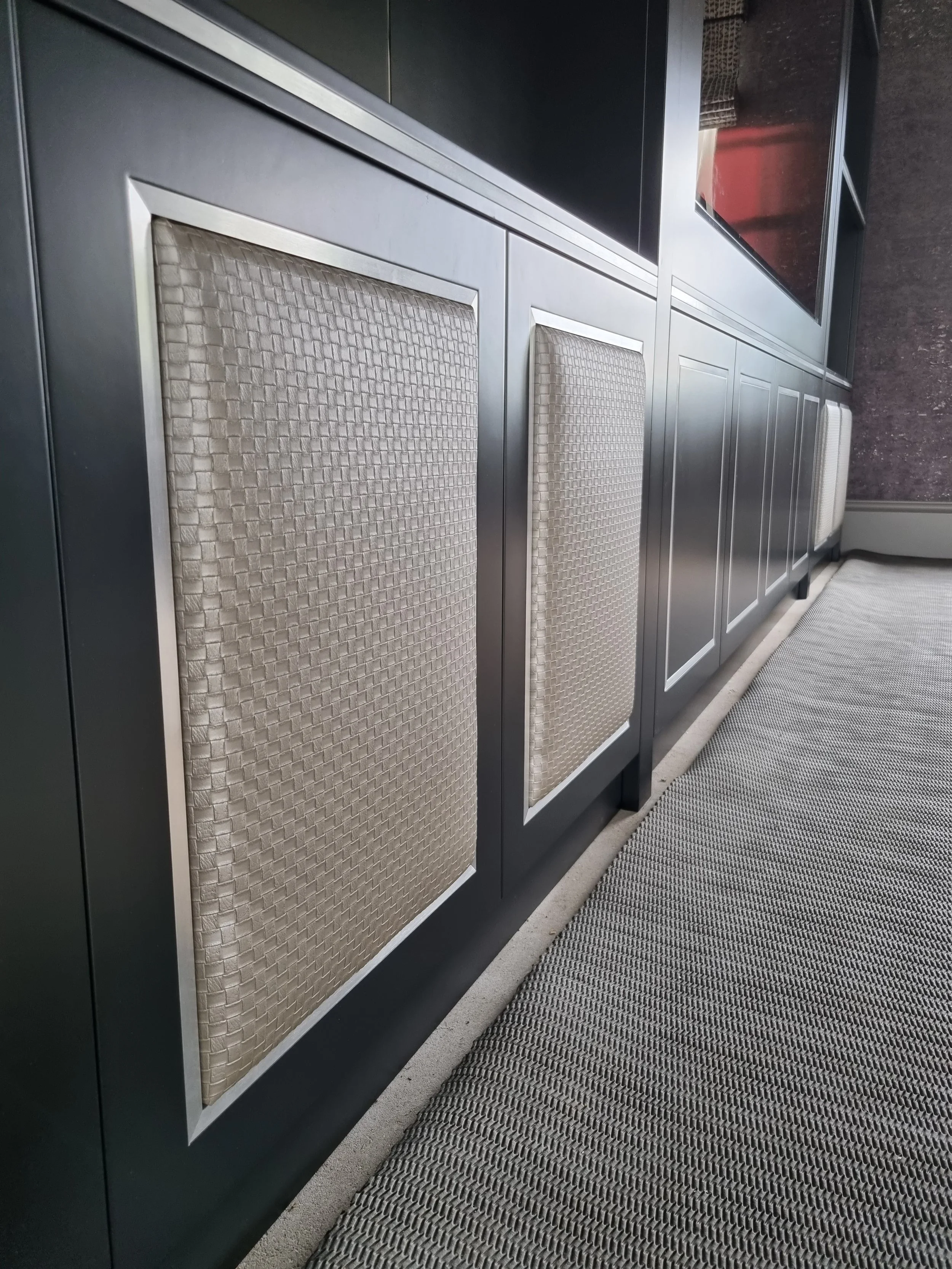 Close-up of a row of air vent grilles with woven fabric covers on a modern black cabinet, adjacent to a textured carpeted floor.