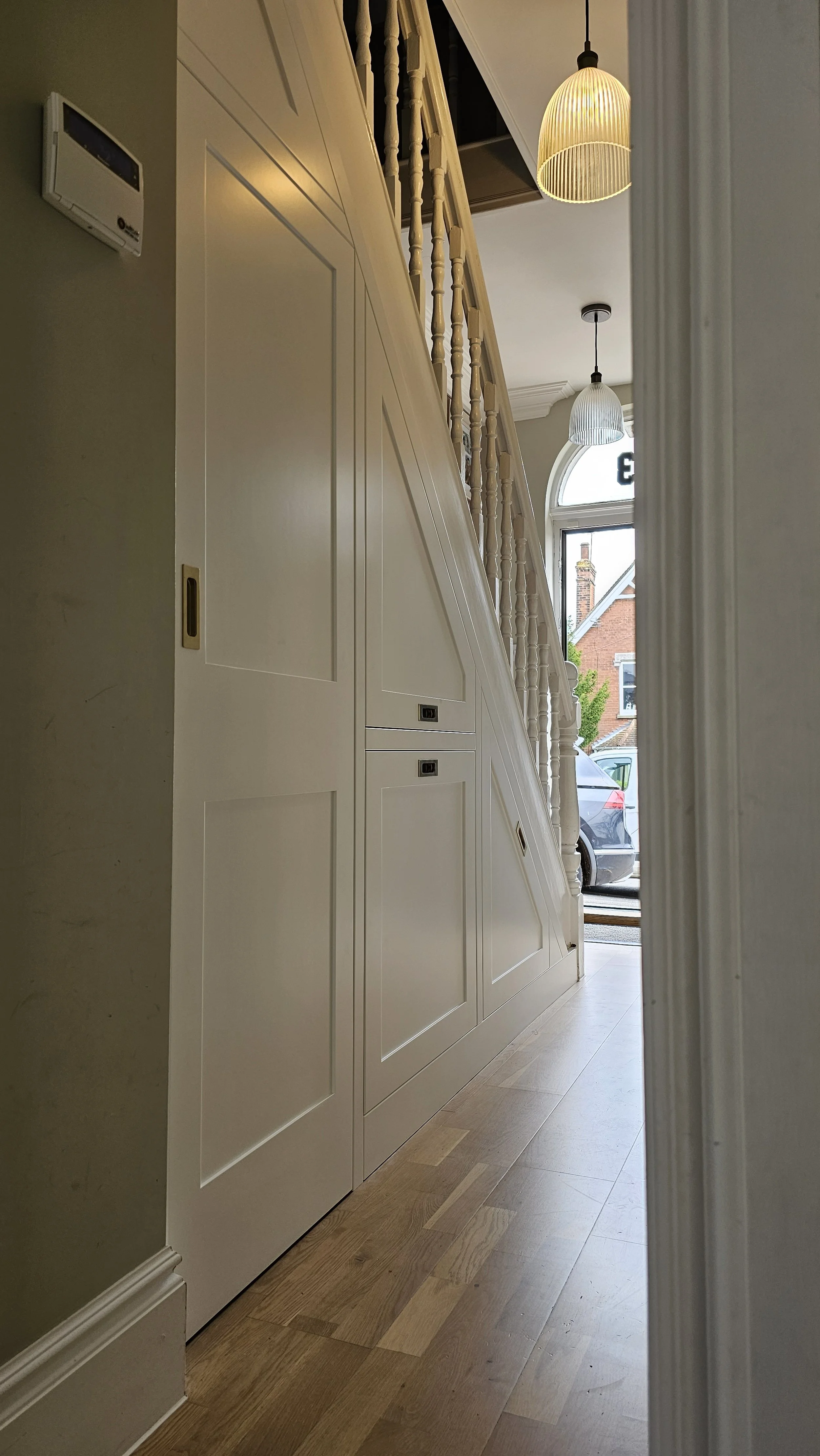 View of a staircase with white paneling and spindled railing, partially visible from a hallway, with hanging pendant lights and a window showing a parked car outside.