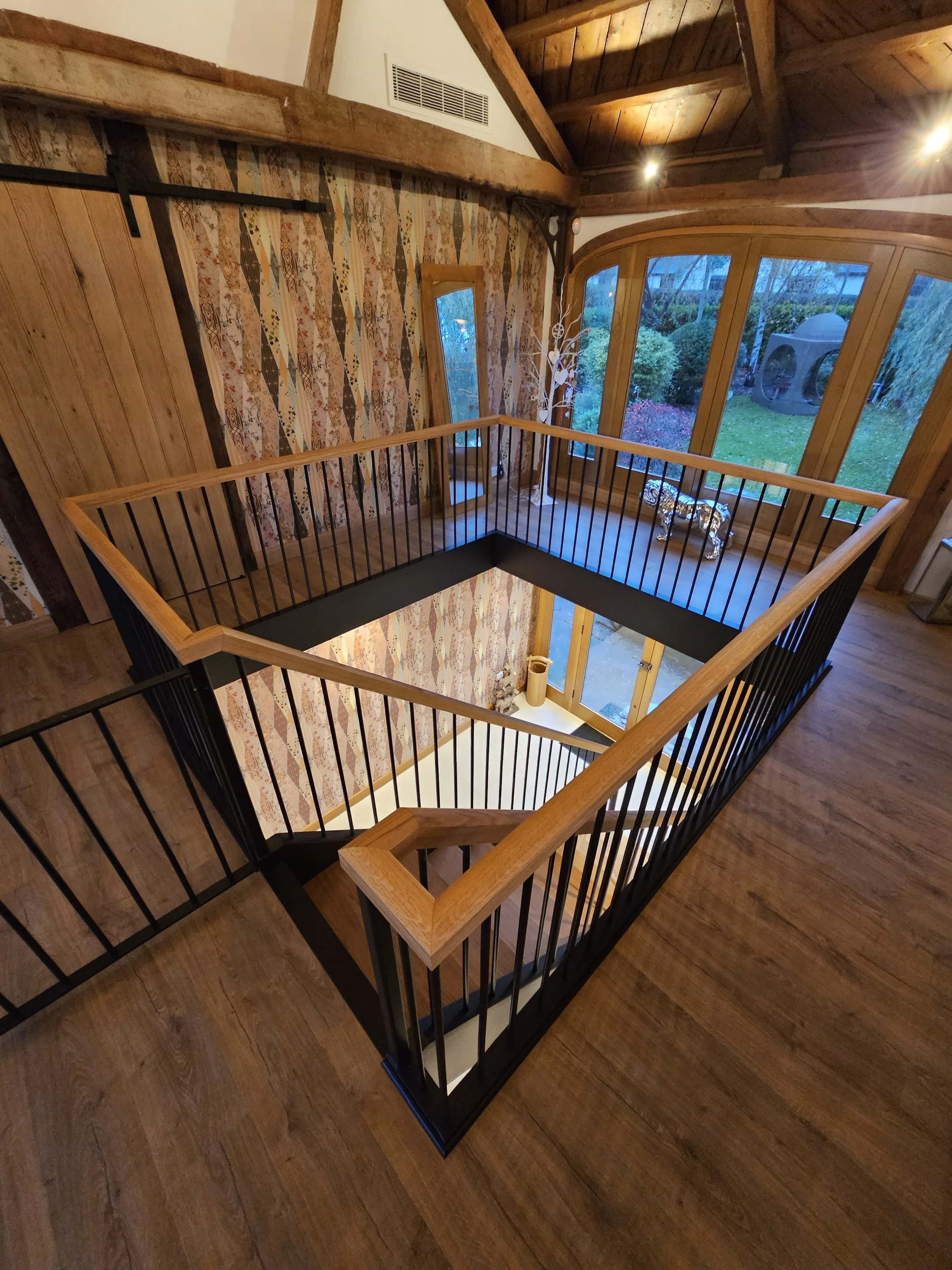 A staircase with black metal railings and wooden handrails surrounding an open area in a house. The upper level has wooden flooring and an interior wall with a patterned design, large windows, and a view of a green backyard with trees and a structure