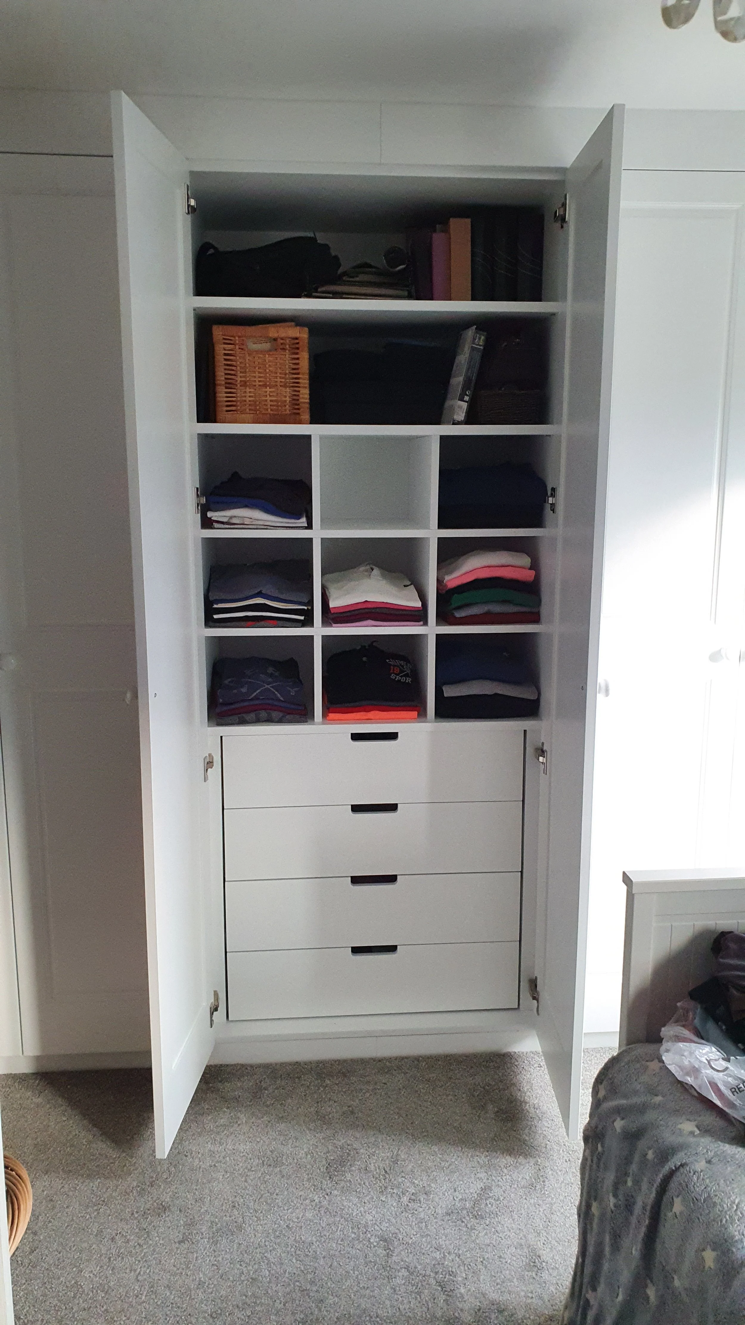 Open white wardrobe with shelves and drawers, containing folded clothes, baskets, books, and bags inside, located in a room with gray carpet and white walls.
