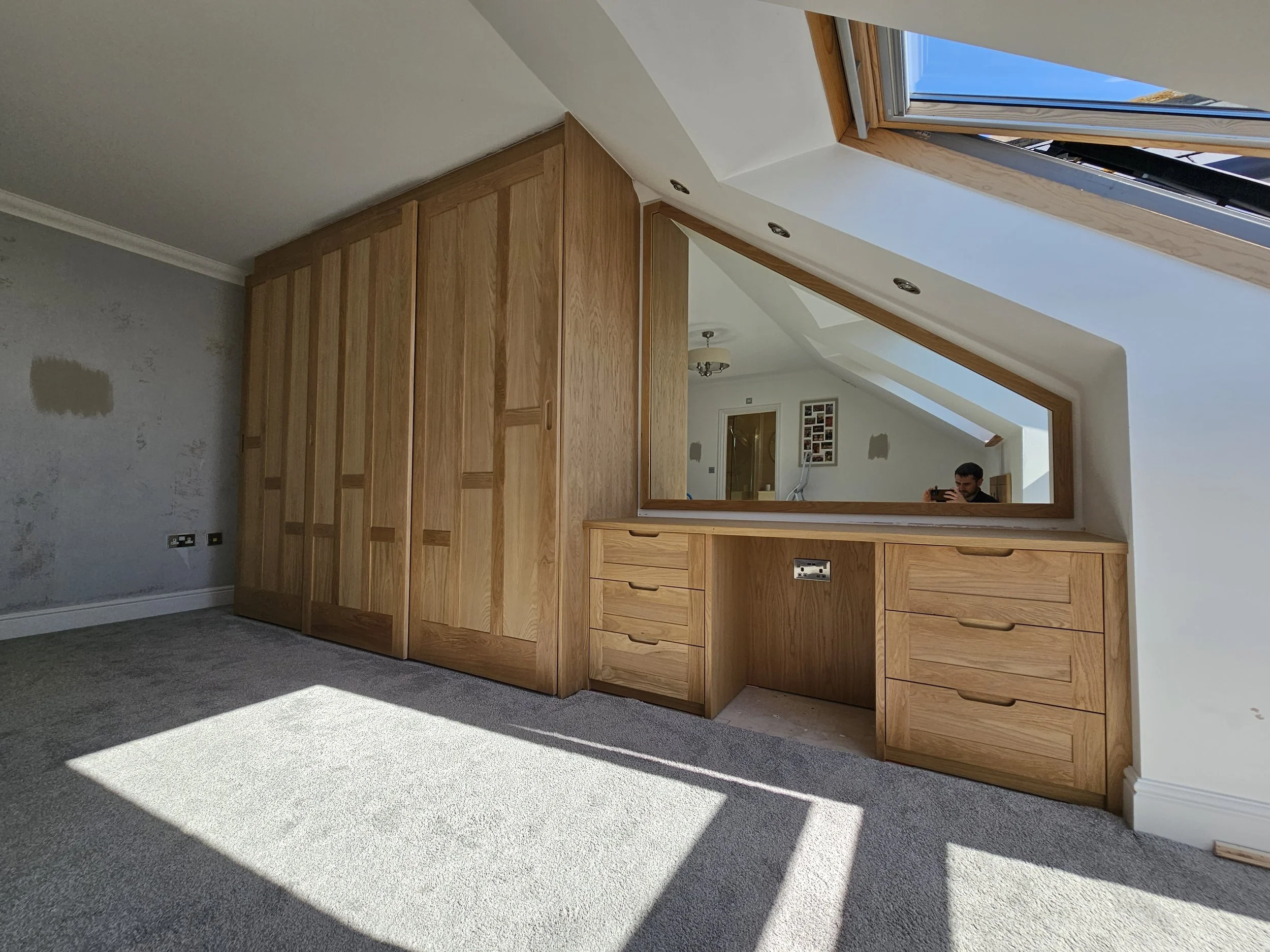Empty room with a large built-in wooden wardrobe, a desk with drawers, and a sloped ceiling with skylight window. The mirror reflects part of a hallway with photos on the wall and a person taking a photo.