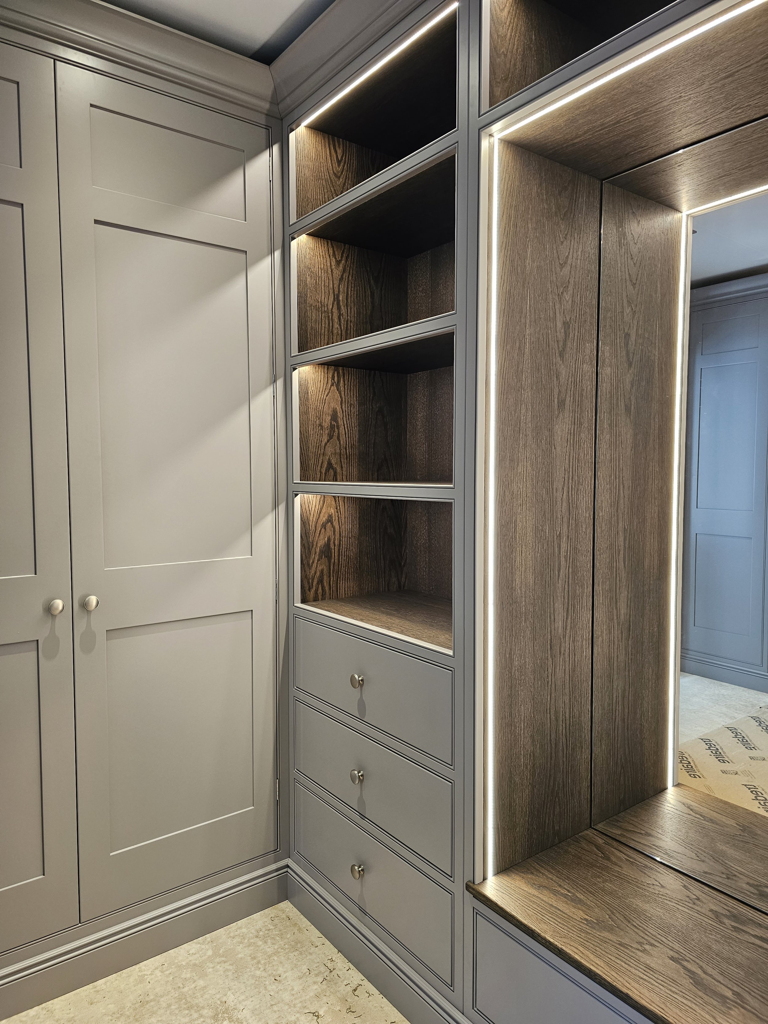 Corner of a modern built-in closet with light grey cabinets, open shelves with warm wood interior, three drawers with small round knobs, and a seating area with wooden top illuminated by integrated lighting.