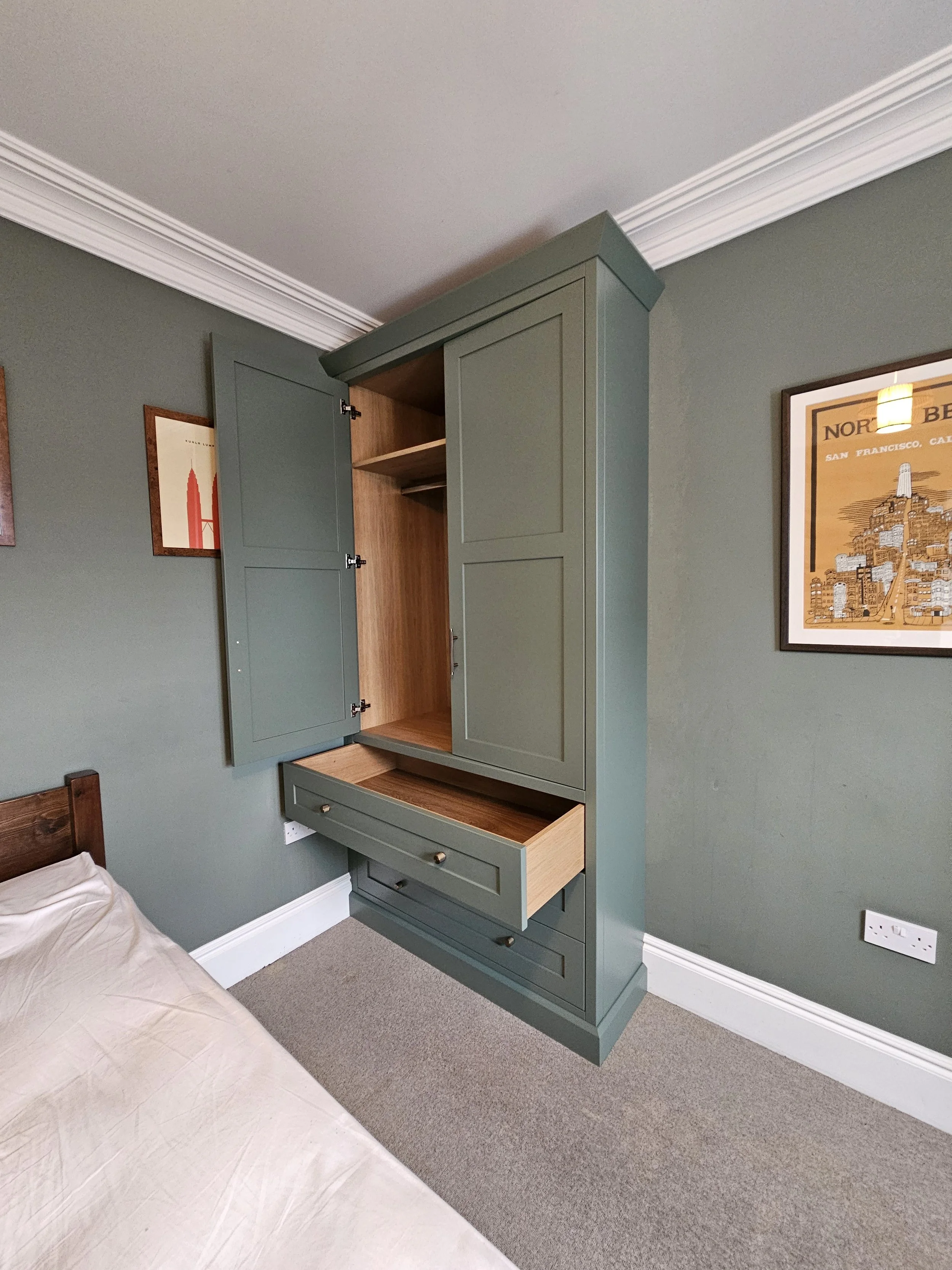 Green wardrobe with open doors and drawers next to a bed in a bedroom with green walls and framed artwork.